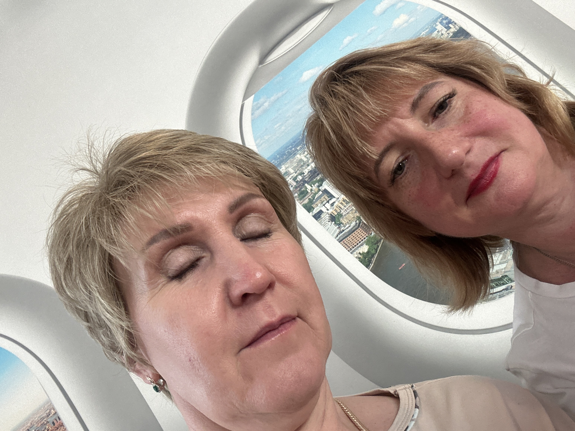 Two women are captured in a close-up, selfie-style photograph, with their faces angled towards the camera. They appear to be inside an airplane cabin, evidenced by the characteristic circular airplane window with a view of a city below. The woman in the foreground has blonde hair, rosy cheeks, and is wearing red lipstick. Her eyes are open, and she is looking towards the camera. The woman in the background, whose face is partially obscured, also has blonde hair and her eyes are closed. She is wearing a light-colored top and a necklace. The city view outside the window shows buildings and a body of water, suggesting a daytime setting. The lighting is bright, casting a soft glow on the subjects. There is no indication of the specific location in Tyumen, Russia, or any other city.