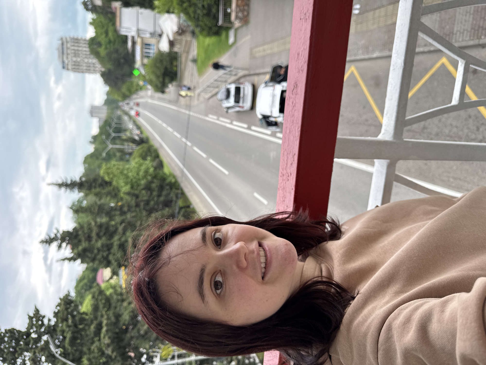 A selfie of a woman in a tan sweatshirt on a bridge in Sochi, Russia. The bridge has a red railing and white supports, with yellow no-parking zig-zags painted on the road beneath. A wide, tree-lined road stretches into the distance, featuring white cars, a person walking, and tall buildings. The environment includes greenery, trees, and a cloudy sky.