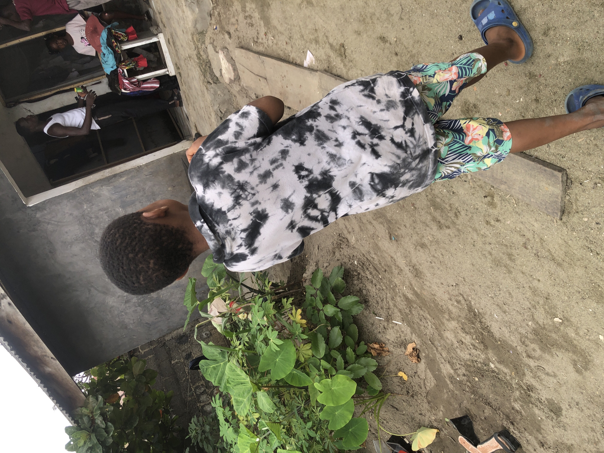 In Ado, Nigeria, a young boy, seen from behind and from the waist down, stands on a dirt ground in front of a simple, unfinished building. He is wearing a tie-dye t-shirt with black and white patterns and colorful, tropical-print shorts. He has blue Croc-style sandals on his feet. To his left, lush green foliage with large leaves grows from the sandy soil. In the background, an adult male in a white tank top and dark pants stands in a doorway, holding a green object. Beside him, a young girl sits on a wooden bench with colorful clothes piled on it. The building appears to be made of concrete blocks and has a corrugated metal roof. The overall scene suggests a rural or semi-urban setting in Nigeria.