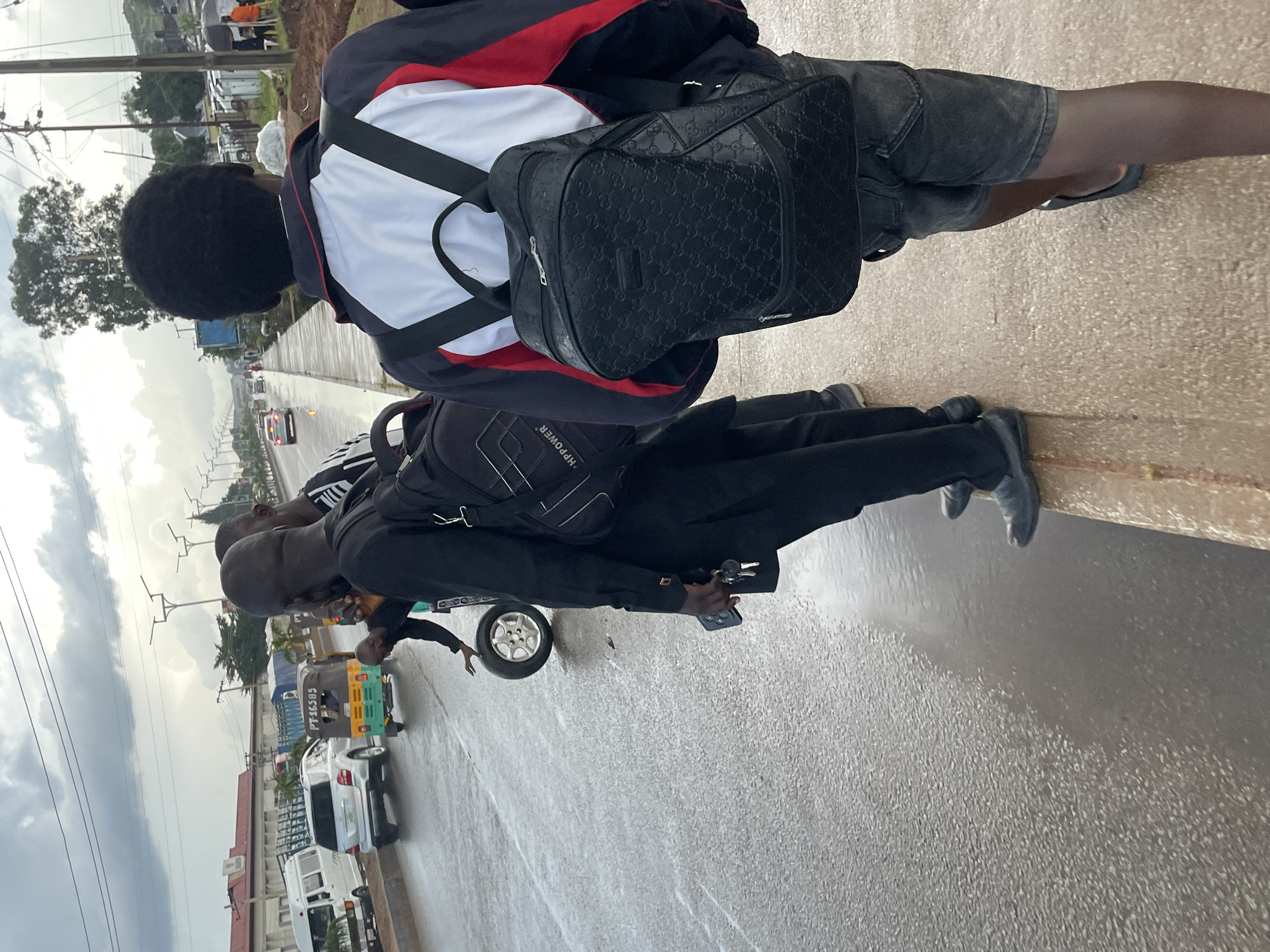 In Barakin Lerawi, Nigeria, three men stand on a wet street next to a sidewalk. The man in the foreground on the right is viewed from behind, wearing a dark jacket with white and red accents and dark denim shorts, carrying a patterned black backpack. To his left, a man in a black outfit and black backpack stands next to a loose tire on the wet asphalt. He holds keys and a phone in his right hand. Behind him, another man in a black shirt and white striped shorts is partially visible. Cars and a trishaw are visible on the street in the background. The sky is overcast.