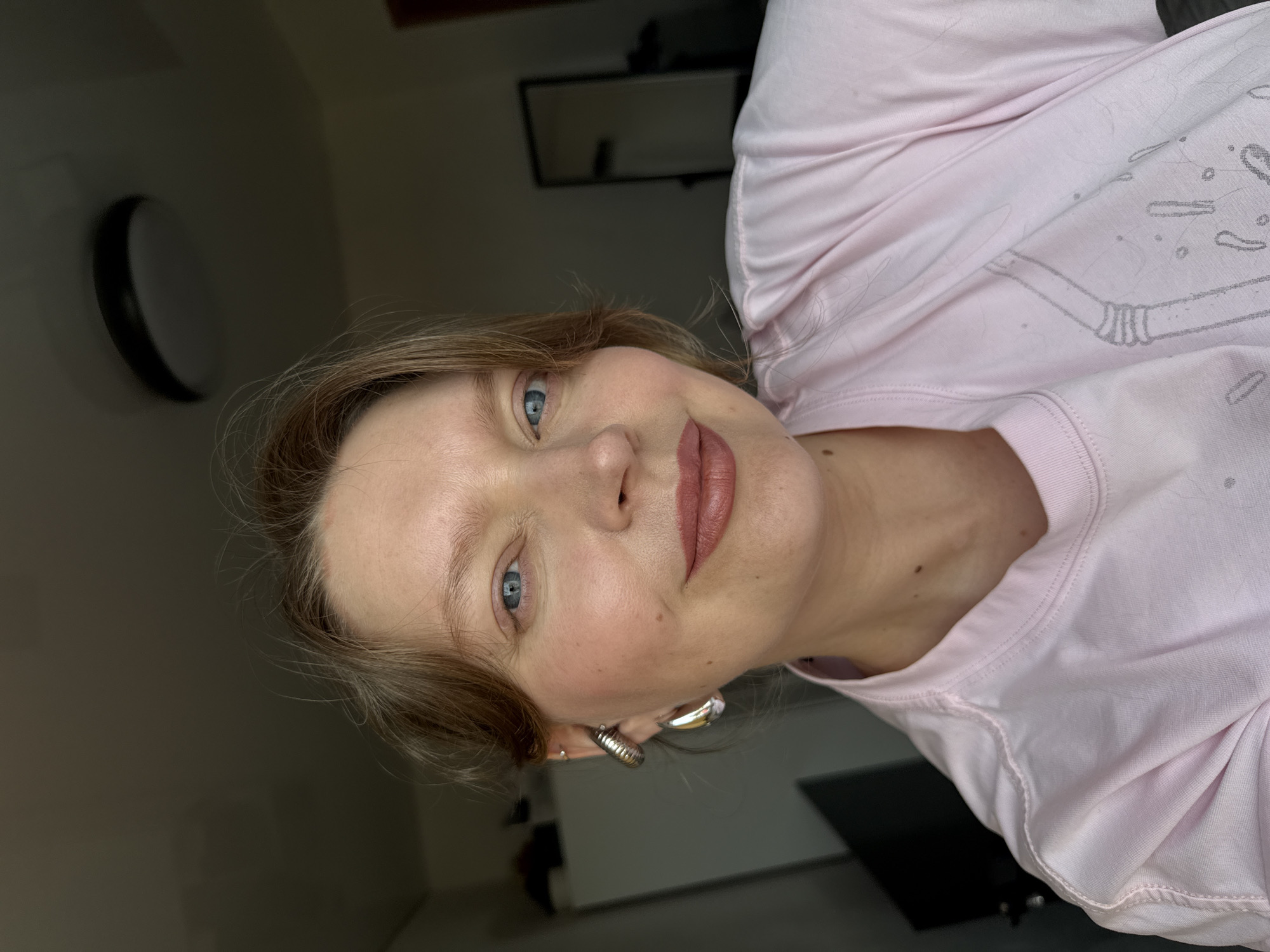 A fair-skinned woman with blue eyes and light brown hair, wearing a light pink shirt, is seen in a close-up shot. She has a mole on her neck and is wearing silver hoop earrings. The background includes a black and white wall-mounted light fixture and a framed mirror, suggesting an indoor environment. The location is not discernible from the image.