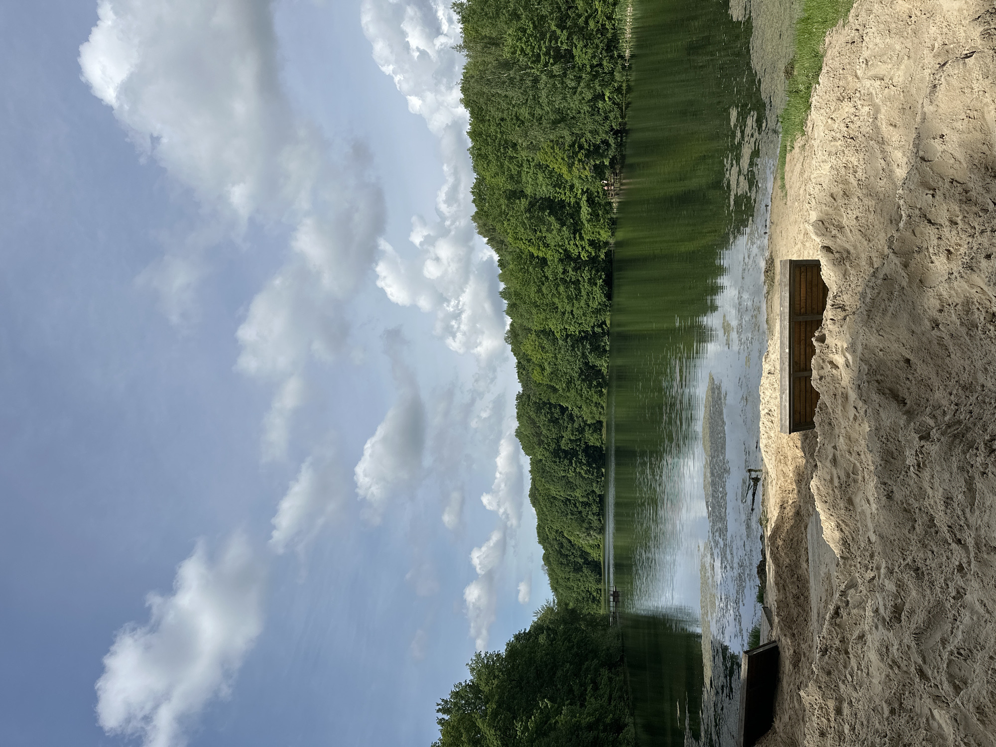 The image shows a scene at the Third Lake of the Swiss Lakes in Nizhny Novgorod, Russia. The view is slightly skewed, showing a body of water (the lake) with a forested shoreline on the opposite side. The water reflects the trees and some of the sky. The sky itself is partly cloudy with a light blue color. In the foreground, there is a sandy area that appears to be a beach or shore, with a small wooden structure embedded into the sand. Vegetation is present along the shoreline where the sand meets the water.
