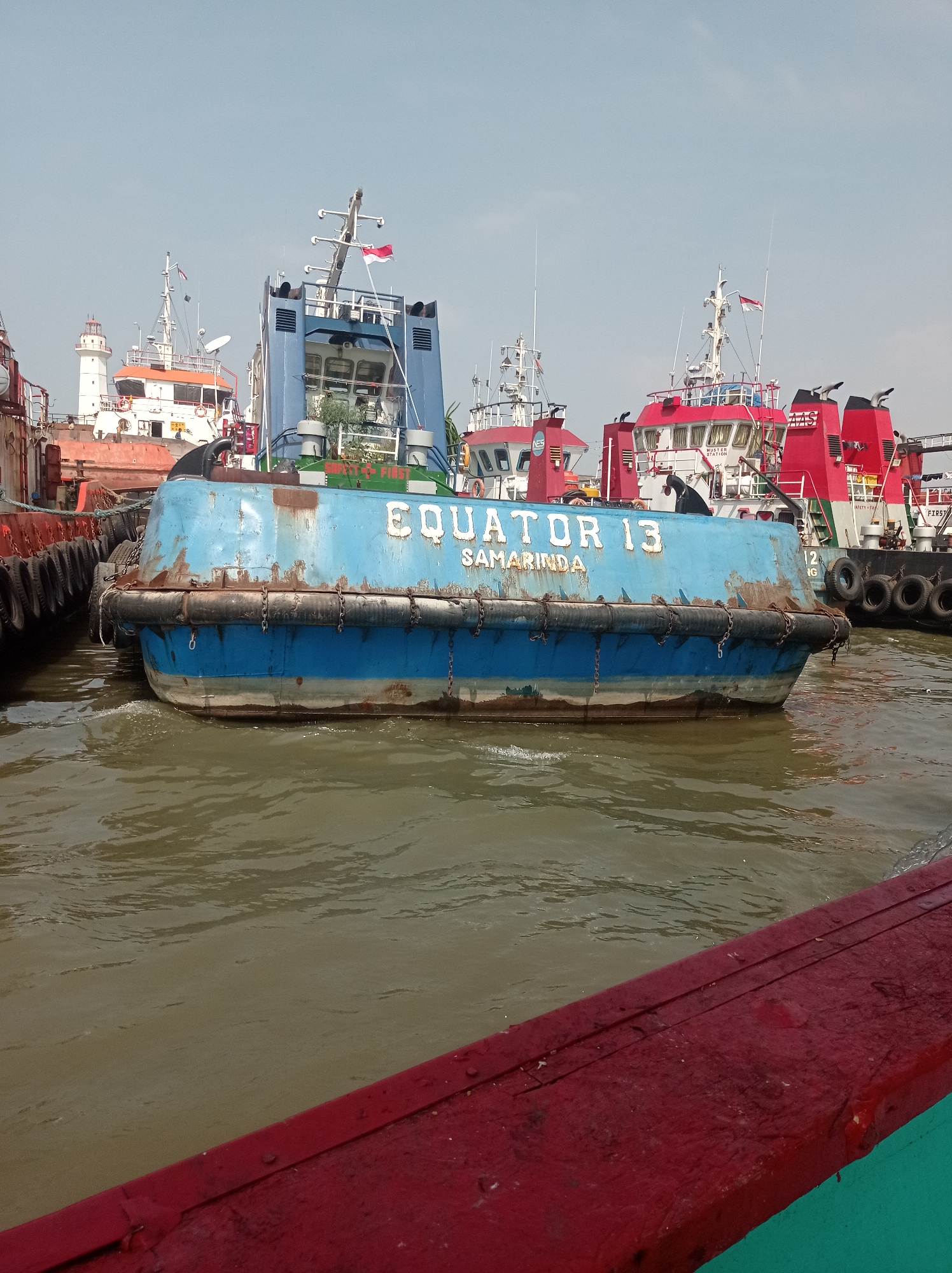 The image shows a harbor with several boats. In the foreground, a blue tugboat named "EQUATOR 13 SAMARINDA" is docked. The boat has a weathered appearance with rust and peeling paint. It is equipped with a black rubber fender and a series of chains along its hull. In the background, several other vessels are visible, including a white lighthouse, a red and white tugboat, and other industrial boats. The water is murky and brown, indicating a river or harbor environment. The sky is clear and light blue. The image appears to have been taken from another boat, as a red railing is visible in the bottom foreground. This scene is likely located in the port of Tegal, Indonesia, given the presence of industrial vessels and the Indonesian flag on one of the boats.