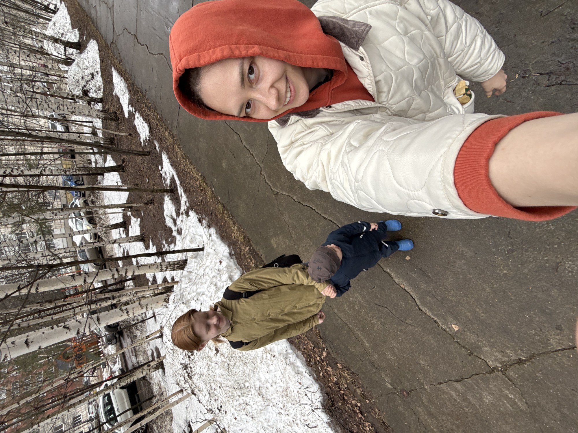 A woman wearing an orange hoodie and white quilted jacket is taking a selfie. Below her and slightly to the left, a younger woman in a khaki jacket holds the hand of a young child wearing a blue snowsuit and brown hat. They are standing on a cracked asphalt walkway with patches of snow along the side. Trees and parked cars are visible in the background. The location is likely Chaikovsky, Russia.