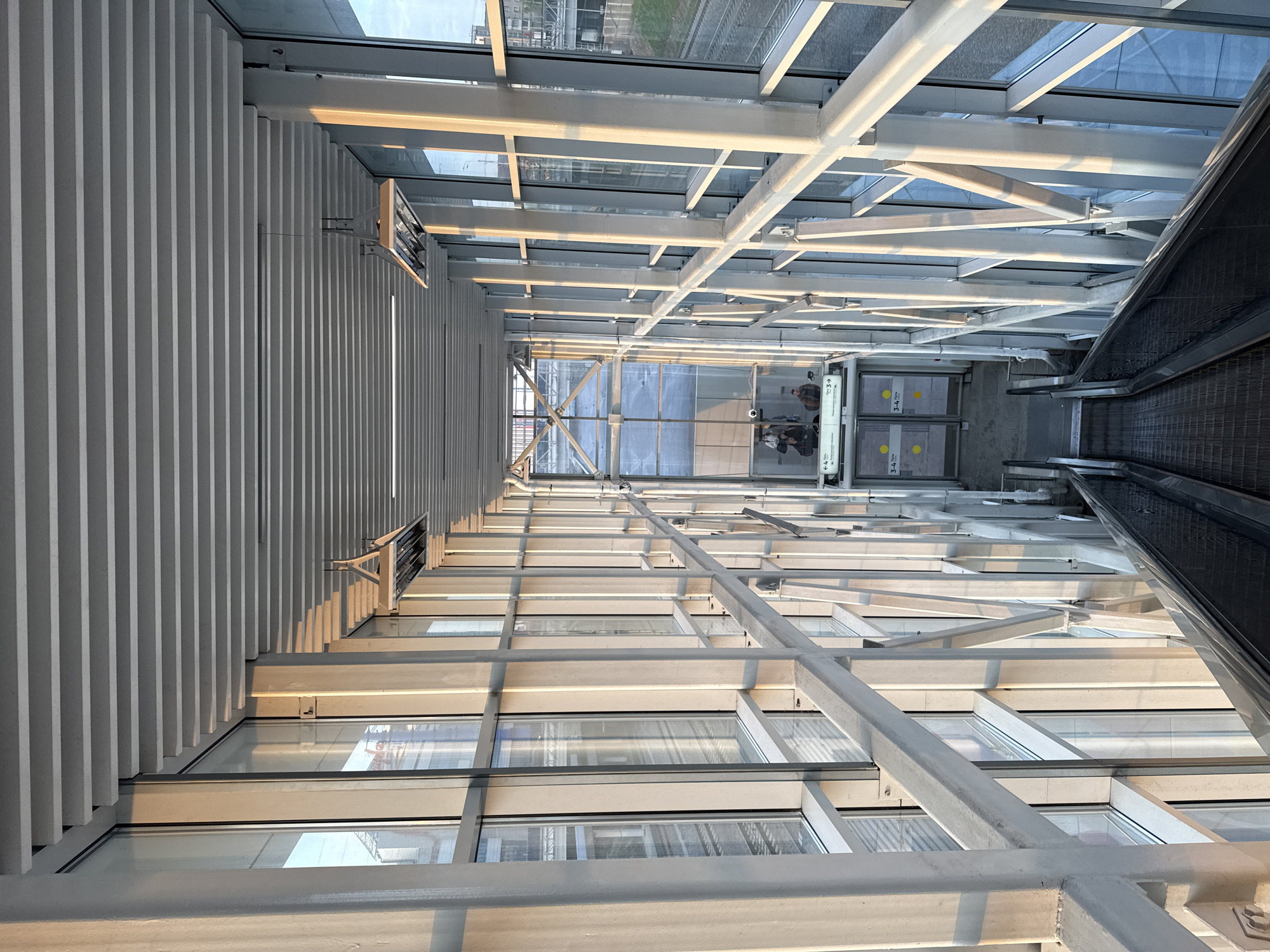 The image shows the interior of a building in Moscow, Russia. The view looks down a long corridor with a metal ceiling, a glass wall, and an escalator. The corridor leads to a doorway with signs. The signs point toward gates 3-4. The architectural elements consist of steel beams, a corrugated metal ceiling and glass. Sunlight reflects throughout the scene.