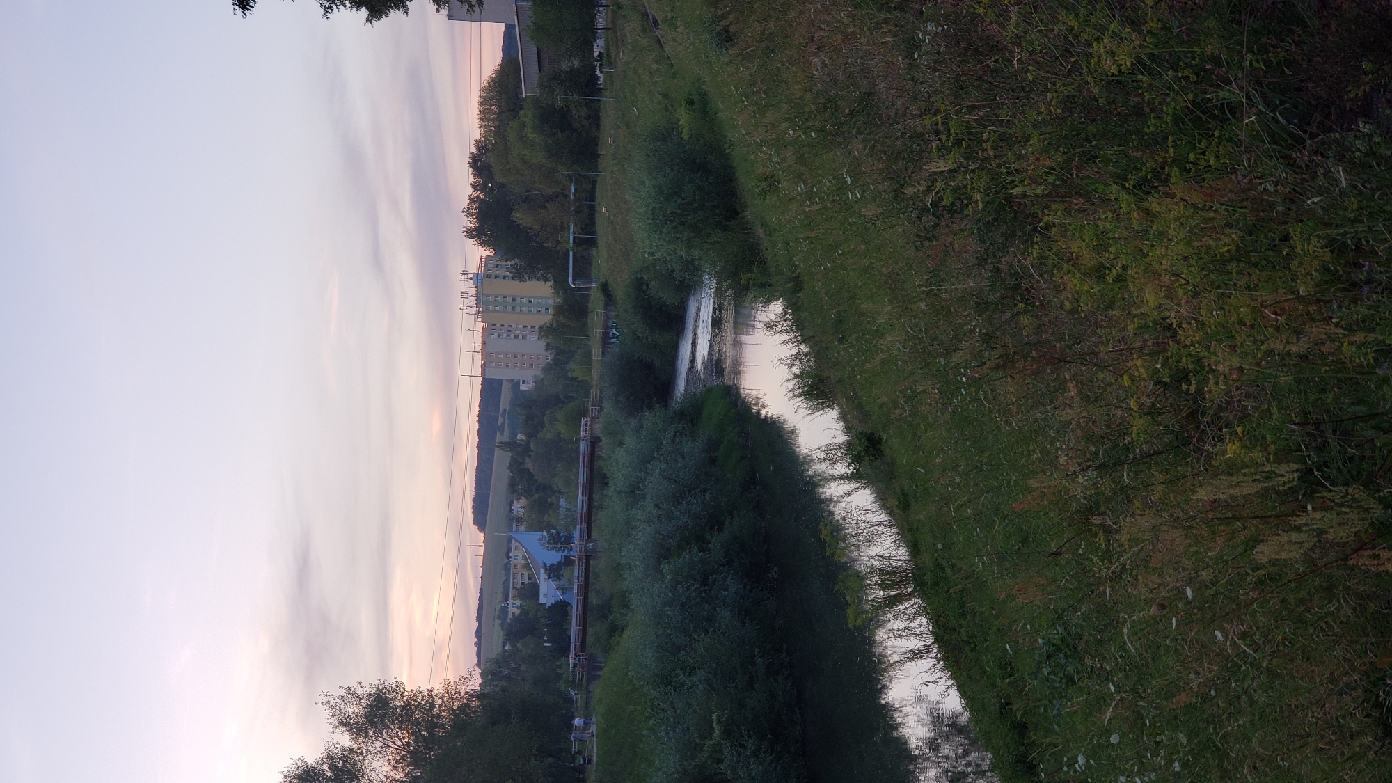 The image depicts a dusk scene in Prešov, Slovakia, likely near the Torysa river. A narrow river flows through the frame, bordered by lush green vegetation on both banks. The left bank is covered in dense, low-lying bushes and trees, while the right bank features a grassy slope with wildflowers and taller grasses. In the background, several residential buildings are visible, including a taller, multi-story apartment building and another structure with a distinct blue architectural design. A bridge spans the river in the mid-ground, with two individuals standing on the pathway near it. The sky above is a soft gradient of pale pink and lavender, indicating the fading light of sunset. The overall atmosphere is serene and tranquil.