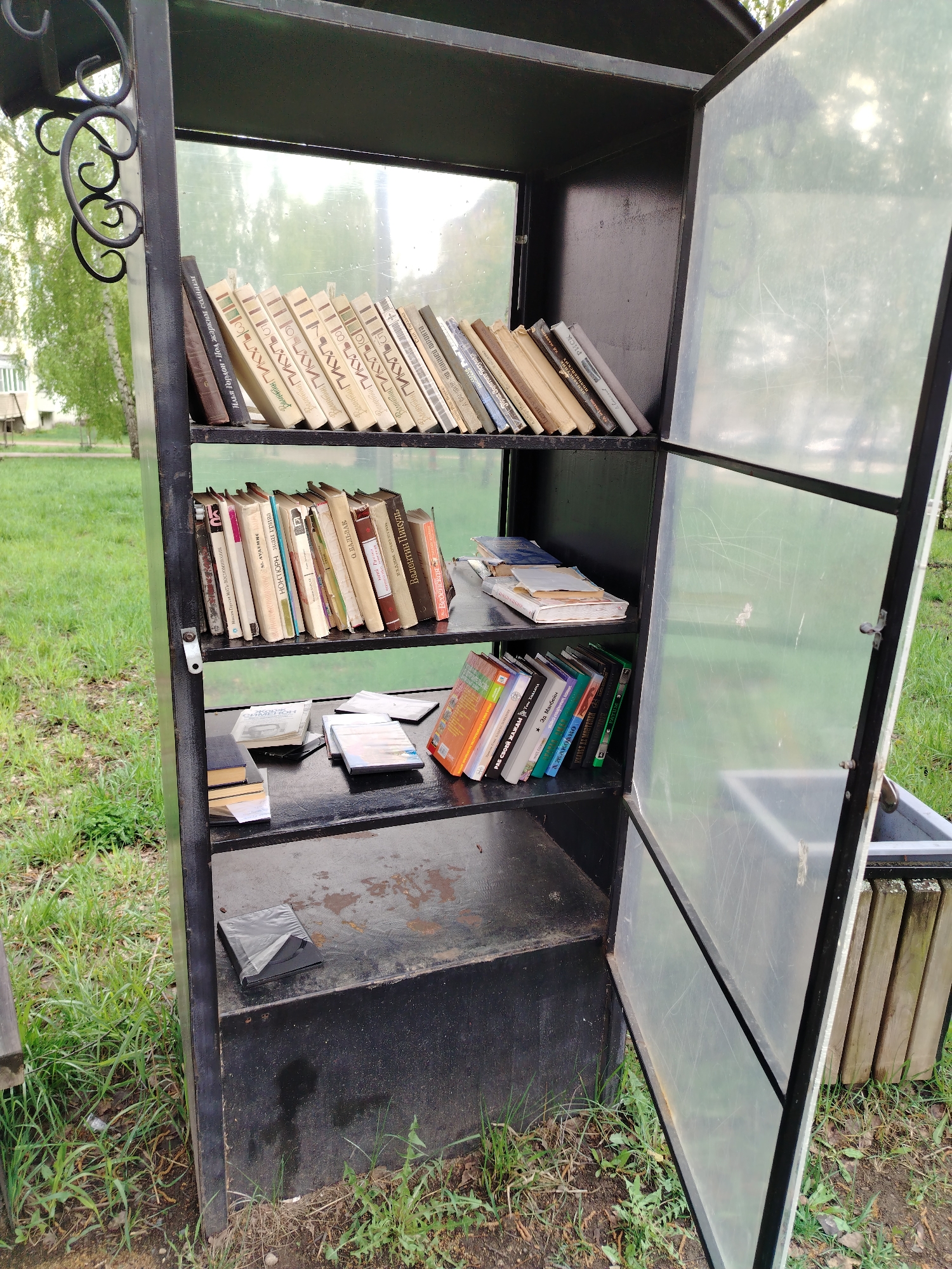 The image captures an outdoor book exchange cabinet in Noviy, Russia. The cabinet is a black metal structure with shelves filled with books. The top shelf contains mainly light-colored books, while the middle shelf has a mix of light and dark-colored books. The bottom shelf contains a mix of books and other items, including what appear to be CDs or DVDs. The cabinet has a frosted glass door that is ajar, revealing the contents. The books appear to be arranged in a haphazard manner, suggesting that people frequently take and leave books. The background shows a grassy area with trees, suggesting a park or public space. The image suggests a community-driven initiative to promote reading and book sharing in Noviy, Russia.