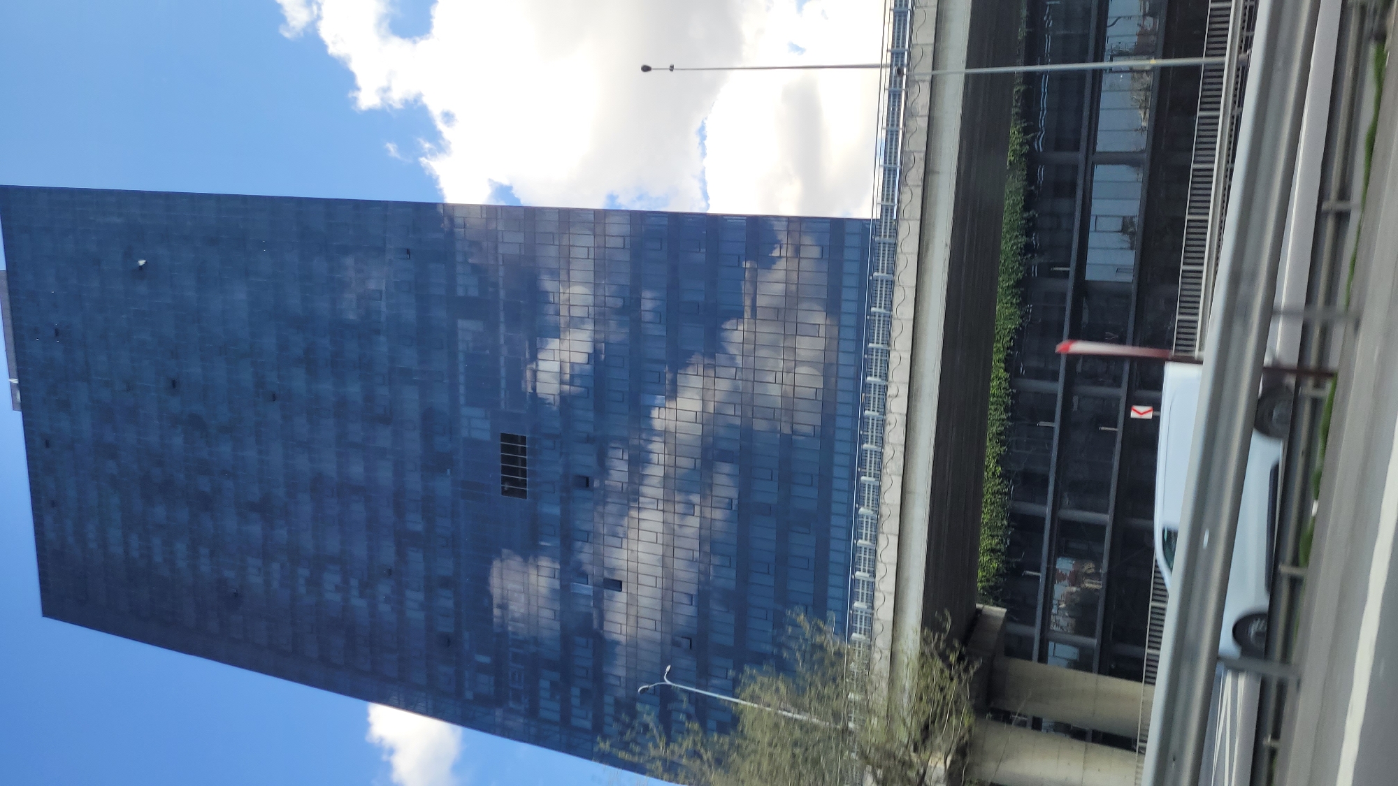 A tall, blue skyscraper with a mirrored facade dominates the left side of the frame, reflecting white clouds in a blue sky. To the right, a concrete overpass structure runs parallel to the building. Green foliage is visible on the side of the overpass closest to the skyscraper. A white van is partially visible in the foreground, partially obscured by a gray metal barrier. The scene is located in Bağcılar, Turkey. A streetlight is also visible.