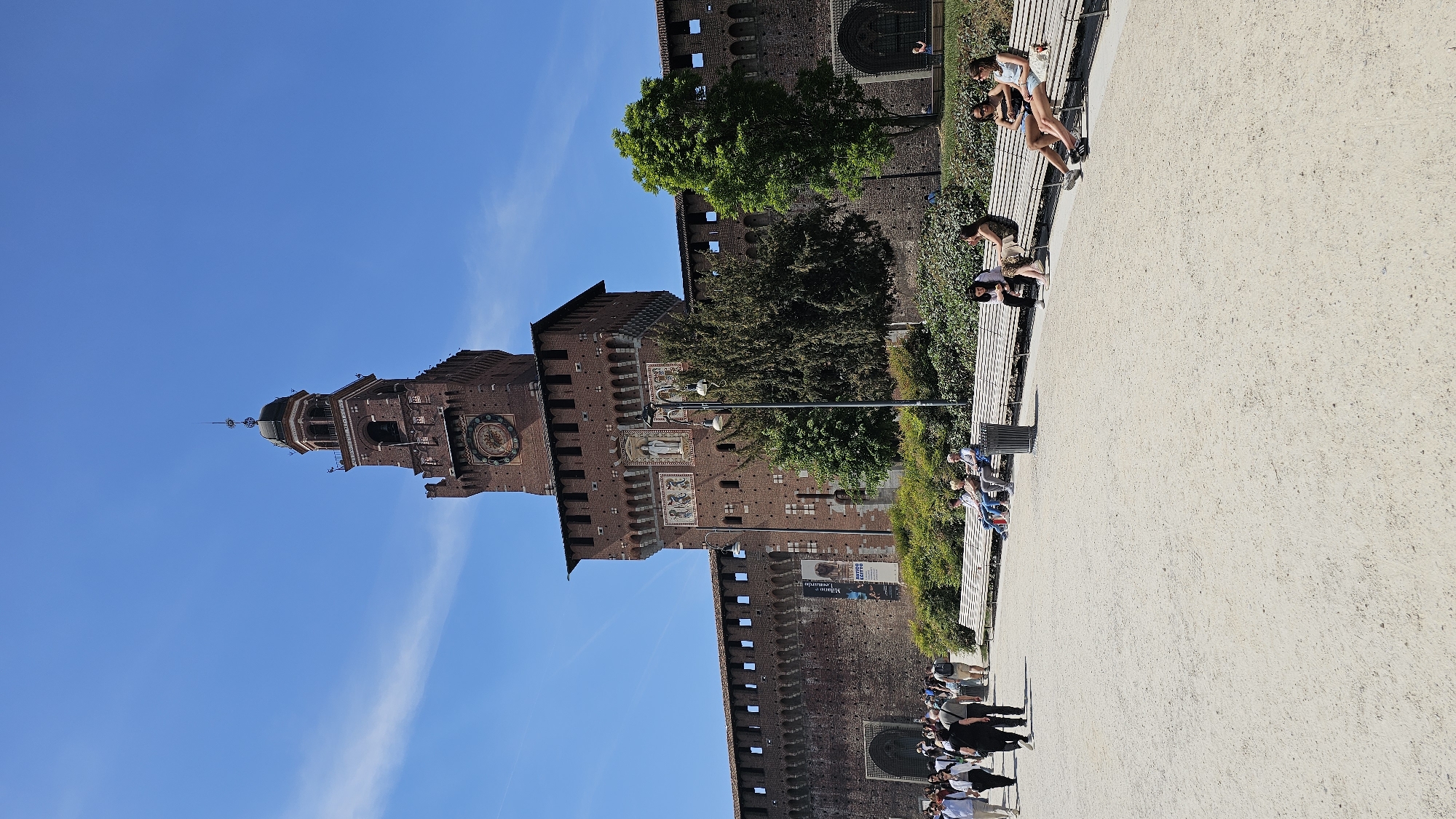 The image is a view of the Sforza Castle (Castello Sforzesco) in Milan, Italy. The castle's tower is visible against a clear blue sky. A long wall of the castle runs along the side, adorned with architectural details and small windows. A line of trees and greenery separates the wall from a pedestrian walkway. People are sitting on benches along the walkway, facing the castle. The ground is covered in light-colored gravel. Some people are seen walking along the path.