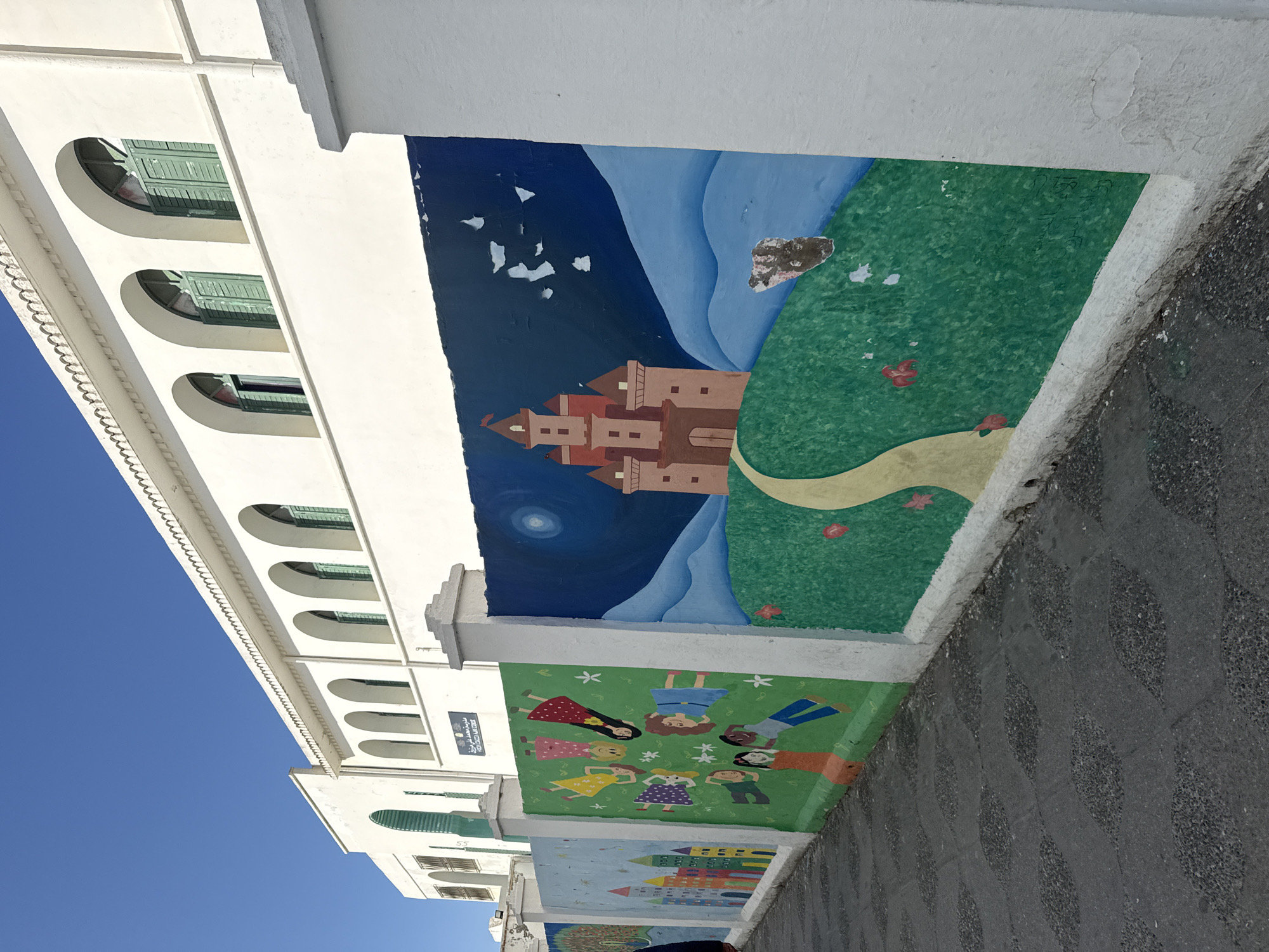 The image shows a street scene in Asilah, Morocco. A white building with arched windows is visible on the left under a bright blue sky. Along the base of the building is a painted mural. The mural is divided into sections. The first section depicts children lying in a circle on green grass. The second section shows a painted castle at night. The foreground shows a sidewalk made of a textured grey material.