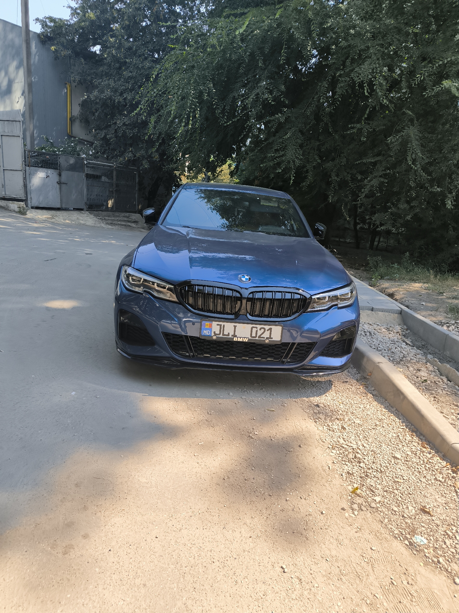 A blue BMW sedan is parked on an asphalt street in Chișinău, Moldova, in front of a large tree with dense foliage and some industrial fencing in the background. The car's front end, including its grille, headlights, and license plate, is clearly visible. The license plate reads "MD JLI 021". The street is partially shaded by the tree, and the ground is a mix of asphalt and gravel. The overall scene suggests an urban environment on a sunny day.