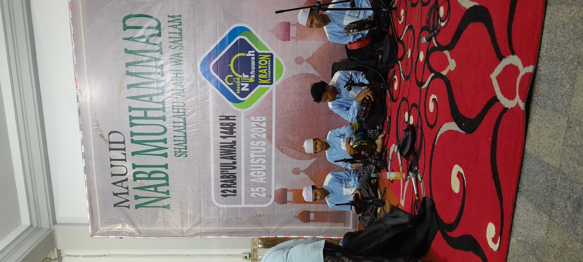 In Bendungan, Indonesia, a group of young boys in blue shirts and white skullcaps are seated on a red carpet with ornate black and white swirling patterns. They are holding and playing hand drums, possibly part of a musical performance. Microphones and cables are visible on the floor in front of them, suggesting a public event. Behind the boys is a large banner with Arabic script and an Islamic crescent moon logo. The banner reads "MAULID NABI MUHAMMAD" and includes dates in both Hijri and Gregorian calendars. The setting appears to be indoors, possibly a mosque or a community hall, decorated with depictions of Islamic architecture like minarets. The overall atmosphere suggests a religious celebration.