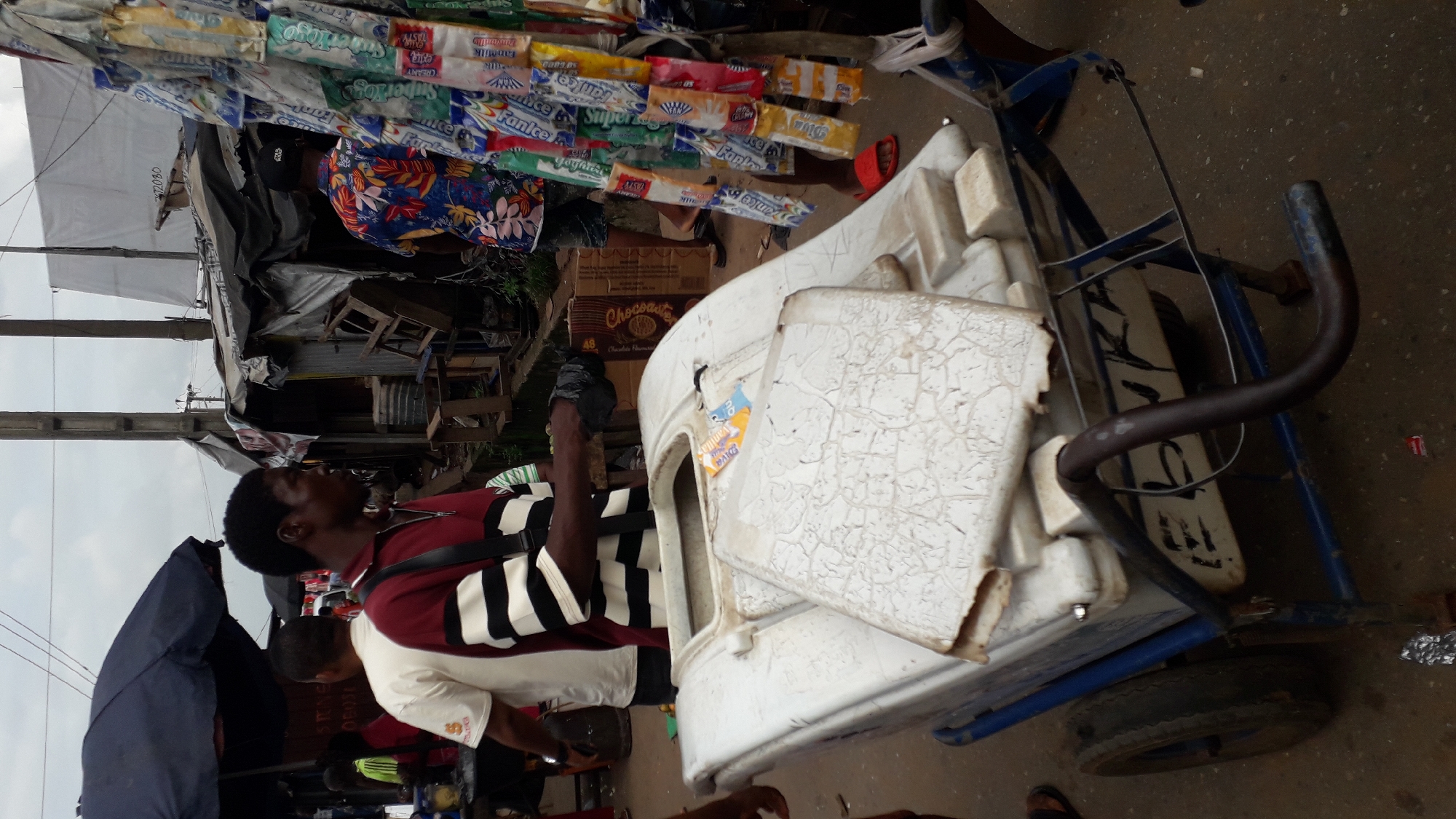 The image depicts an outdoor scene in Nkpor, Nigeria, likely a marketplace. A Black male, wearing a red and black striped shirt and a dark crossbody bag, is standing next to a large white cooler on wheels. He is looking to his right, and his right hand is holding a black plastic bag. To his left is another Black male wearing a white t-shirt. In the background, there are several makeshift stalls with corrugated metal roofs and tarps. A variety of goods are displayed on racks, including packaged snacks or sweets hanging from a string. The ground is paved with dirt and some debris. The overall impression is one of a busy, informal market environment.