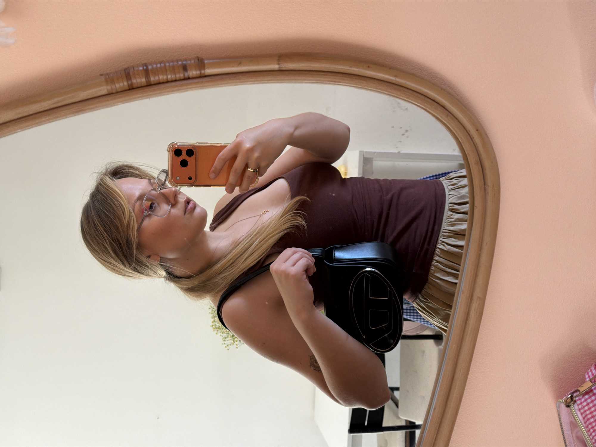 A young woman is taking a mirror selfie. She is wearing glasses, a brown tank top, and a gold skirt. She is holding a smartphone in her right hand and a black bag with a prominent logo on her shoulder. The mirror has a wooden frame. The environment is a room with white walls and a few pieces of furniture visible in the reflection. There is not enough information to confidently say this picture was taken in Tel-Aviv, Israel.