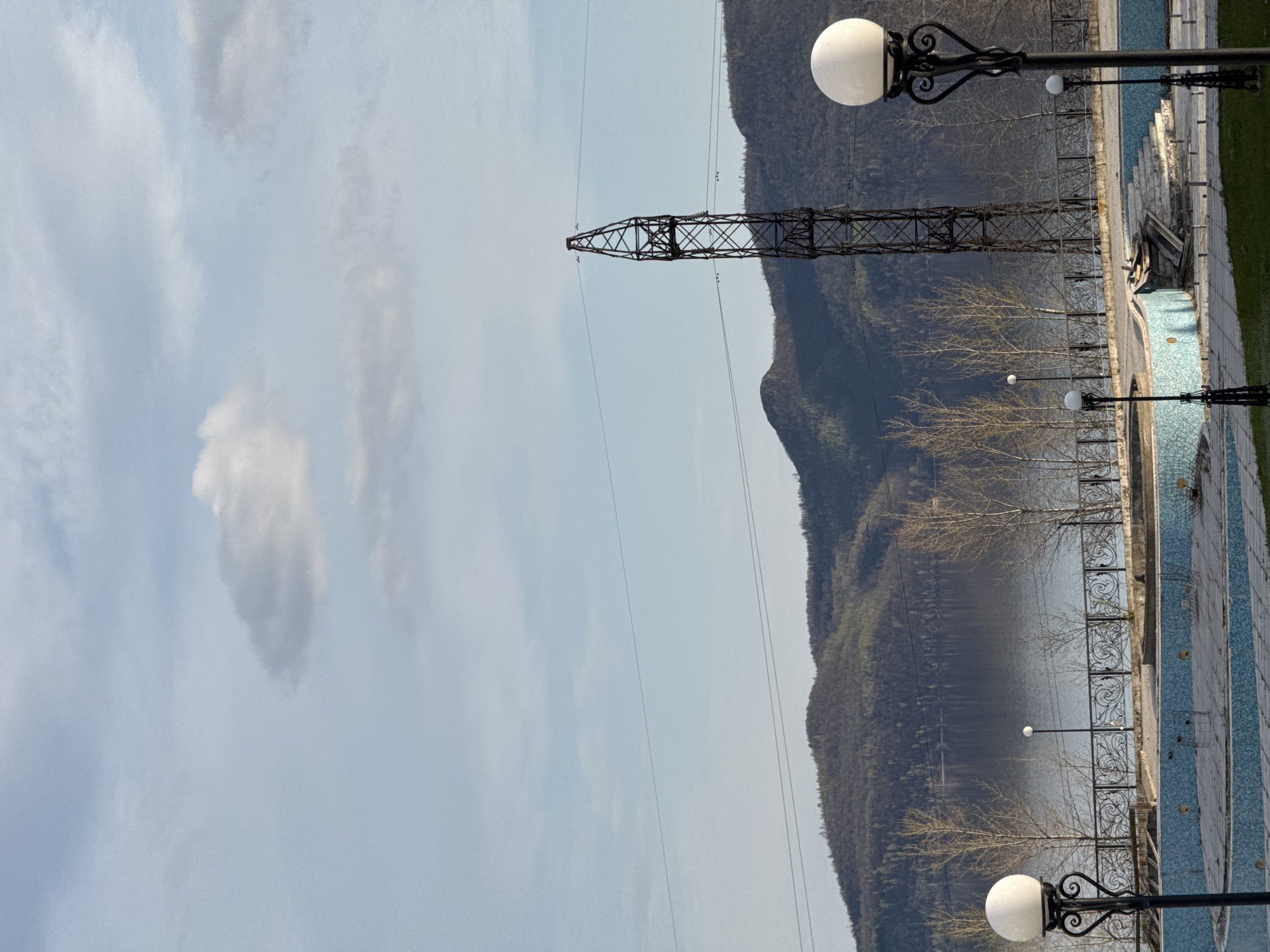 An outdoor landscape image, originally rotated, depicts a serene scene dominated by natural and man-made elements under a partly cloudy sky. In the background, a range of large, tree-covered mountains stretches across the horizon, descending towards a calm body of water, likely a lake or wide river, which reflects the sky and distant slopes.
A prominent, tall power line tower, constructed of dark metal lattice work, stands vertically in the mid-ground towards the left, with thin power lines extending from it across the sky.
In the foreground and mid-ground on the right, there is a paved promenade or walkway running parallel to the water's edge. This area features decorative elements including several ornate black lamp posts with spherical white lights. One such lamp post is clearly visible in the right foreground, and others recede into the distance. A black, scrolled metal fence separates the walkway from the water. The paved surface next to the water appears to be adorned with light blue and white tiles, forming a patterned area that might be a decorative pool or a dried-up water feature. Scattered along the water's edge and promenade are several deciduous trees with bare branches, suggesting late winter or early spring.
The sky above is a light blue with scattered white and light grey clouds. The overall lighting indicates daytime, possibly late morning or early afternoon, with no harsh shadows. No people or specific activities are visible in the tranquil scene. There is no visible text in the image.