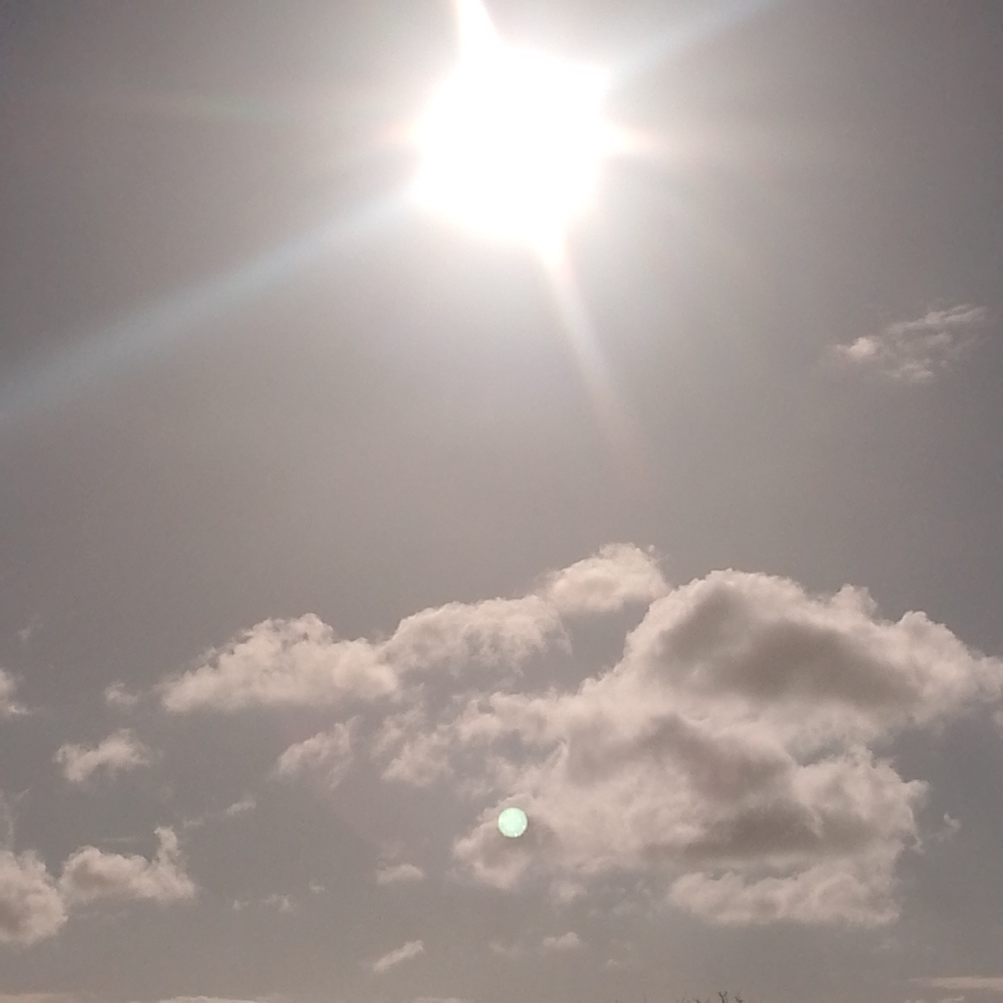 The image shows a daytime sky with the sun prominently visible at the top. The sun's rays extend outwards. Below the sun, there are scattered clouds of varying sizes and shapes. A distinct, spherical object is visible through a break in the clouds; it has a greenish hue. The location, Altagracia, Venezuela, is not explicitly discernible from the image alone, but is inferred from the prompt's instruction. No people are visible.