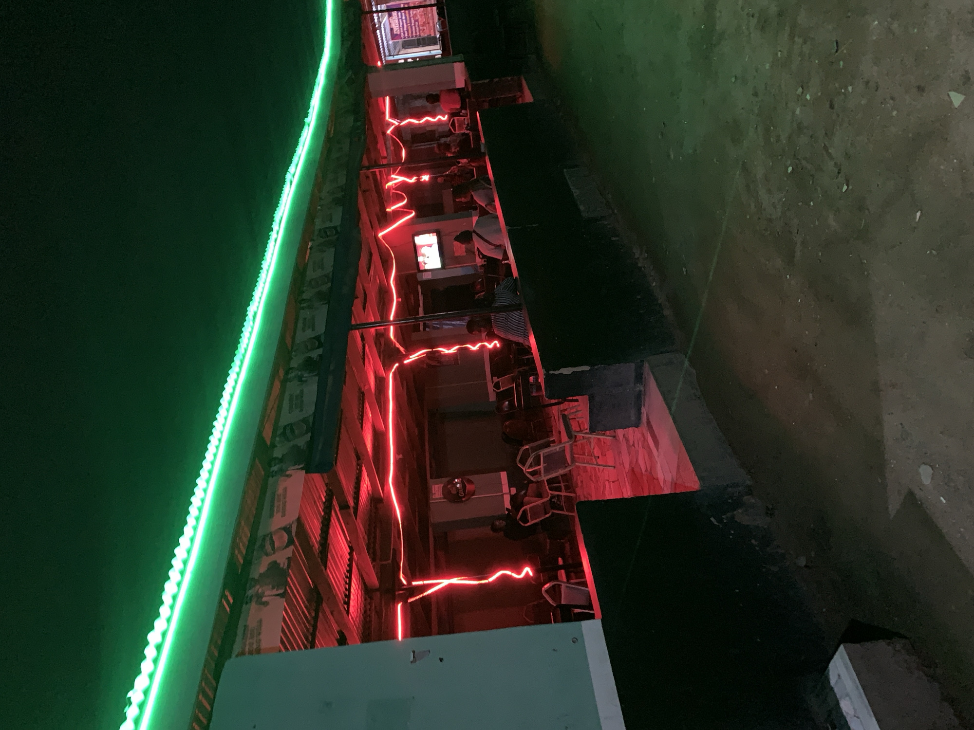 The image shows an outdoor restaurant or bar in the vicinity of the New Stadium in Nigeria. The building is illuminated with green LED strip lights along the roofline and red neon-style lights that zig-zag along the open-air dining area. Patrons are seated at tables and chairs within the red-lit space. A television screen is visible within the restaurant, apparently being watched by some of the patrons. The ground outside the restaurant is dimly lit, suggesting it is nighttime.