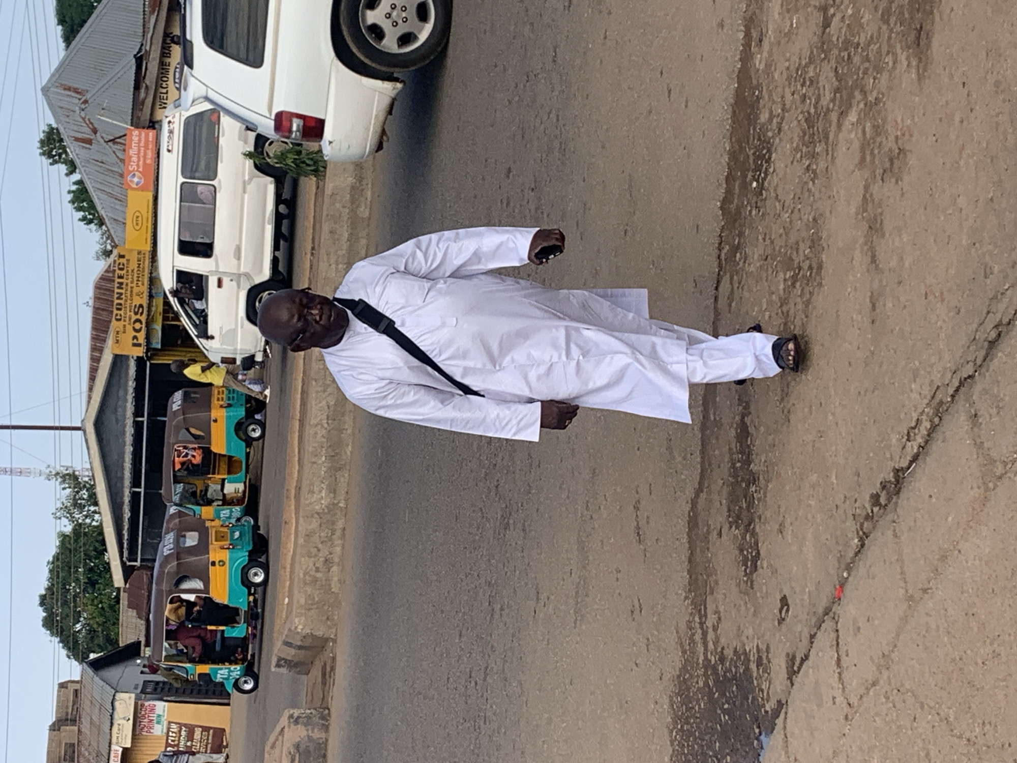 In Gura Top, Nigeria, a man wearing a white robe and black sandals walks across a paved road. He carries a black bag slung across his chest and holds a dark-colored mobile phone in his right hand. In the background, two brightly colored auto-rickshaws are parked on the side of the road, and a white van is also visible. Further in the distance, several buildings line the street, some with signs that read "MTN CONNECT" and "WELCOME BACK." The sky is overcast, suggesting a cloudy day.