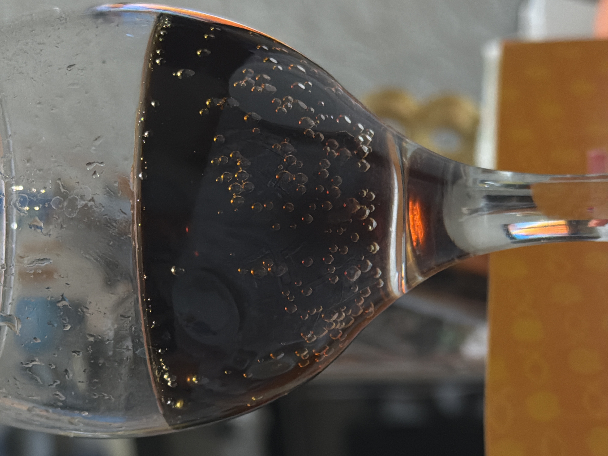 The image displays a close-up, rotated view of a clear glass containing a dark, carbonated beverage, likely a soft drink or beer. The glass is oriented horizontally, with the liquid occupying the bottom portion of the frame in this rotated perspective.
Numerous small, bright bubbles cling to the interior surface of the glass, illuminated by an unseen light source, giving them a golden or amber hue. More bubbles are suspended within the dark liquid, especially near the glass wall. The outside of the glass exhibits condensation and small water droplets, indicating a cold drink.
The background is softly blurred. To the right of the glass, a warm-toned, possibly orange or light brown, rectangular object (such as a box or package) is visible. Other indistinct shapes and colors are present in the deeper background. The setting appears to be indoors. No people or visible text are present. The overall focus is on the textures and reflections of the glass and the effervescence of the drink.