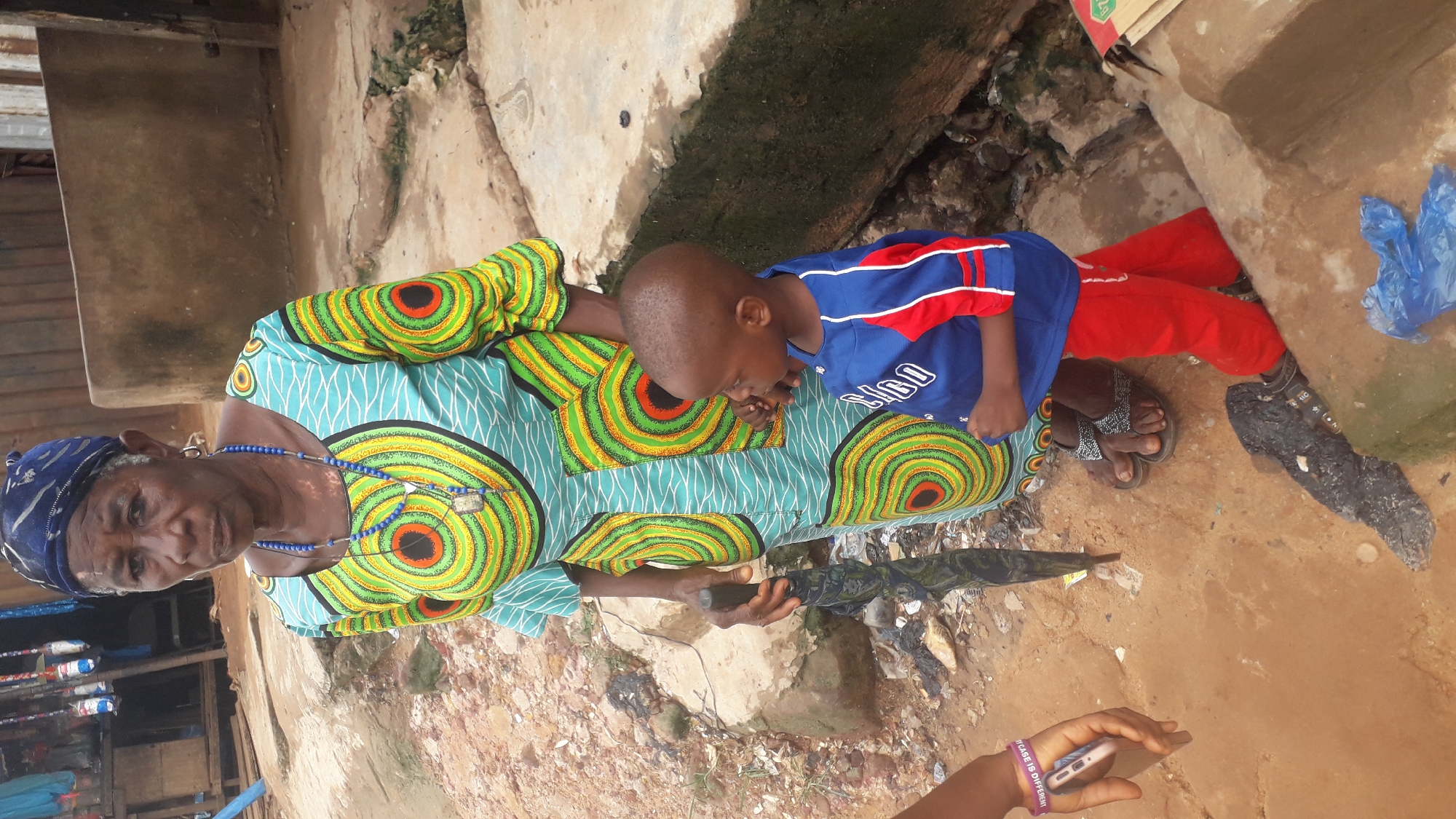 An elderly woman and a young child are positioned in an outdoor environment in Nkpor, Nigeria. The woman is wearing a colorful patterned top with a blue headscarf and beaded necklaces. The child is wearing a blue and red shirt with red pants. The woman is holding an umbrella. The background consists of buildings, concrete structures, and what appears to be litter and debris. A hand holding a phone is visible in the foreground.
