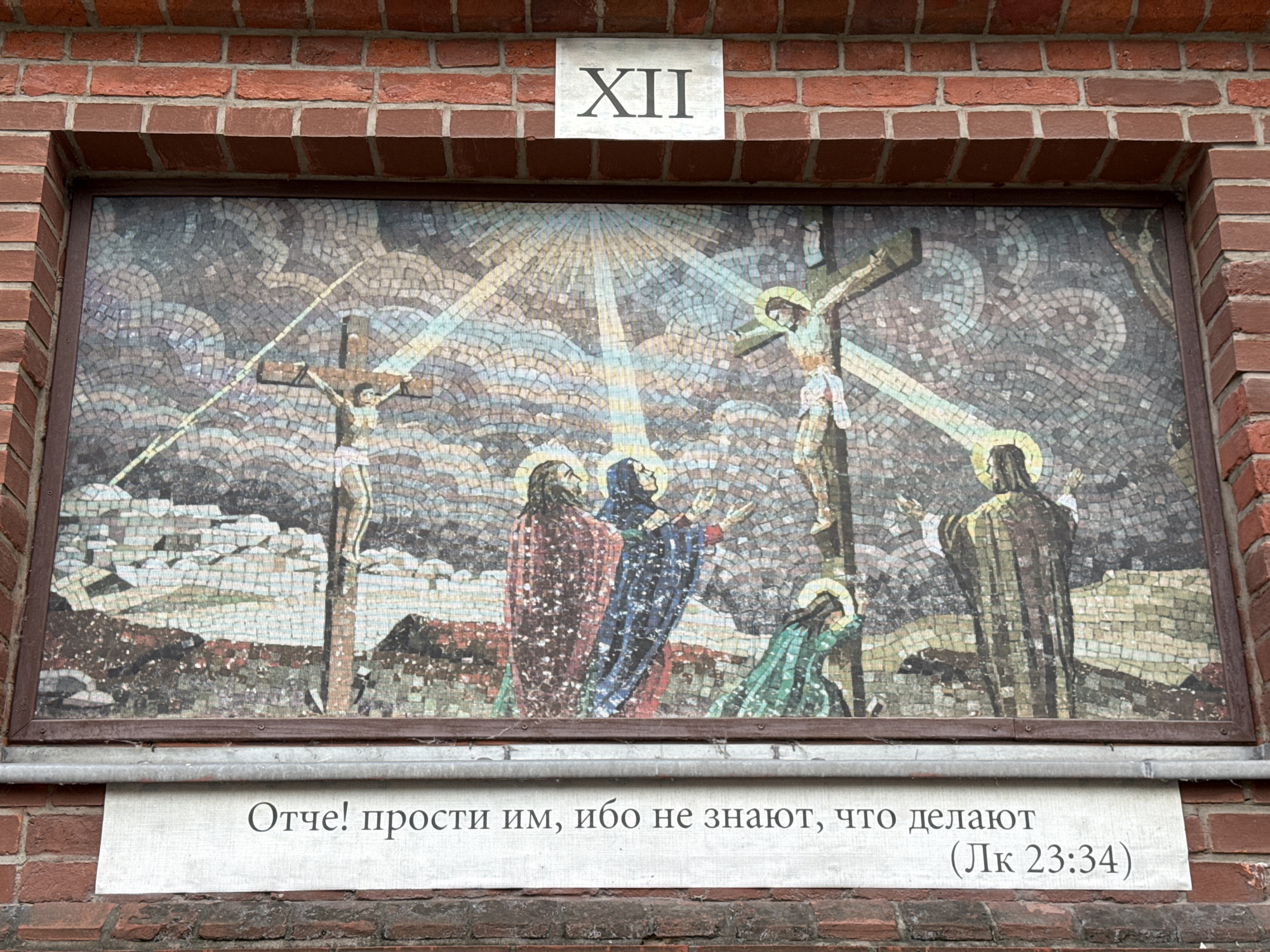 The image shows a mosaic depicting the crucifixion of Jesus Christ, accompanied by two other individuals on crosses. Below the mosaic, a text in Russian reads "Отче! прости им, ибо не знают, что делают" which translates to "Father! forgive them, for they know not what they do." This quote is attributed to Luke 23:34, as indicated by "(Лк 23:34)" below the text. The mosaic is mounted on a brick wall, and above it, a plaque displays the Roman numeral "XII". This artwork is located at the Church of the Resurrection of the Word in Moscow, Russia, as part of its Stations of the Cross.