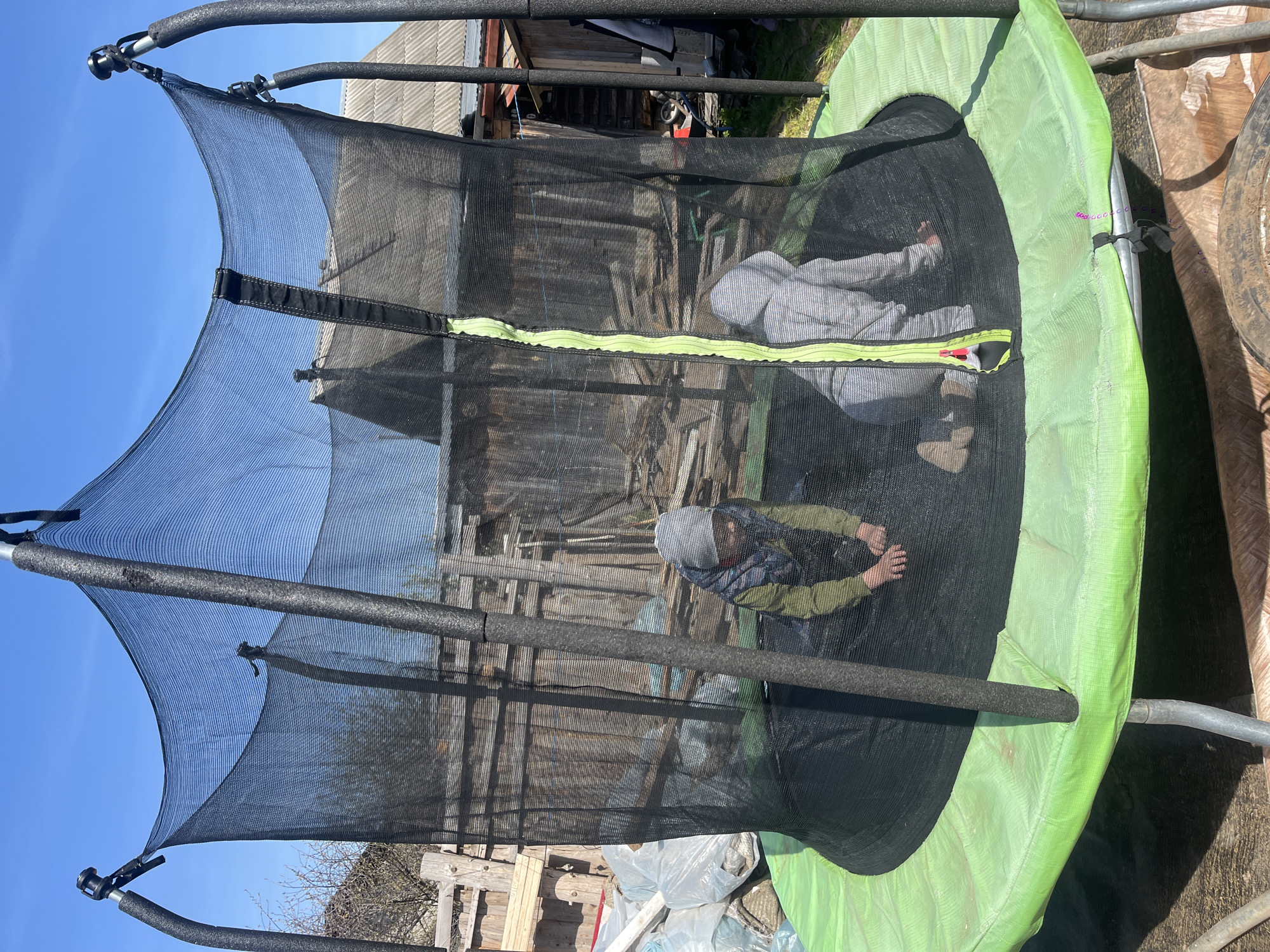 Two children are inside a trampoline enclosed by a black safety net. The trampoline has a green padded rim. Outside the trampoline, there is a cluttered yard with wooden planks, debris, and the corner of a building. The sky is blue. The location is outdoors. The precise location in the city of Двигатель, Russia, cannot be determined from the image.