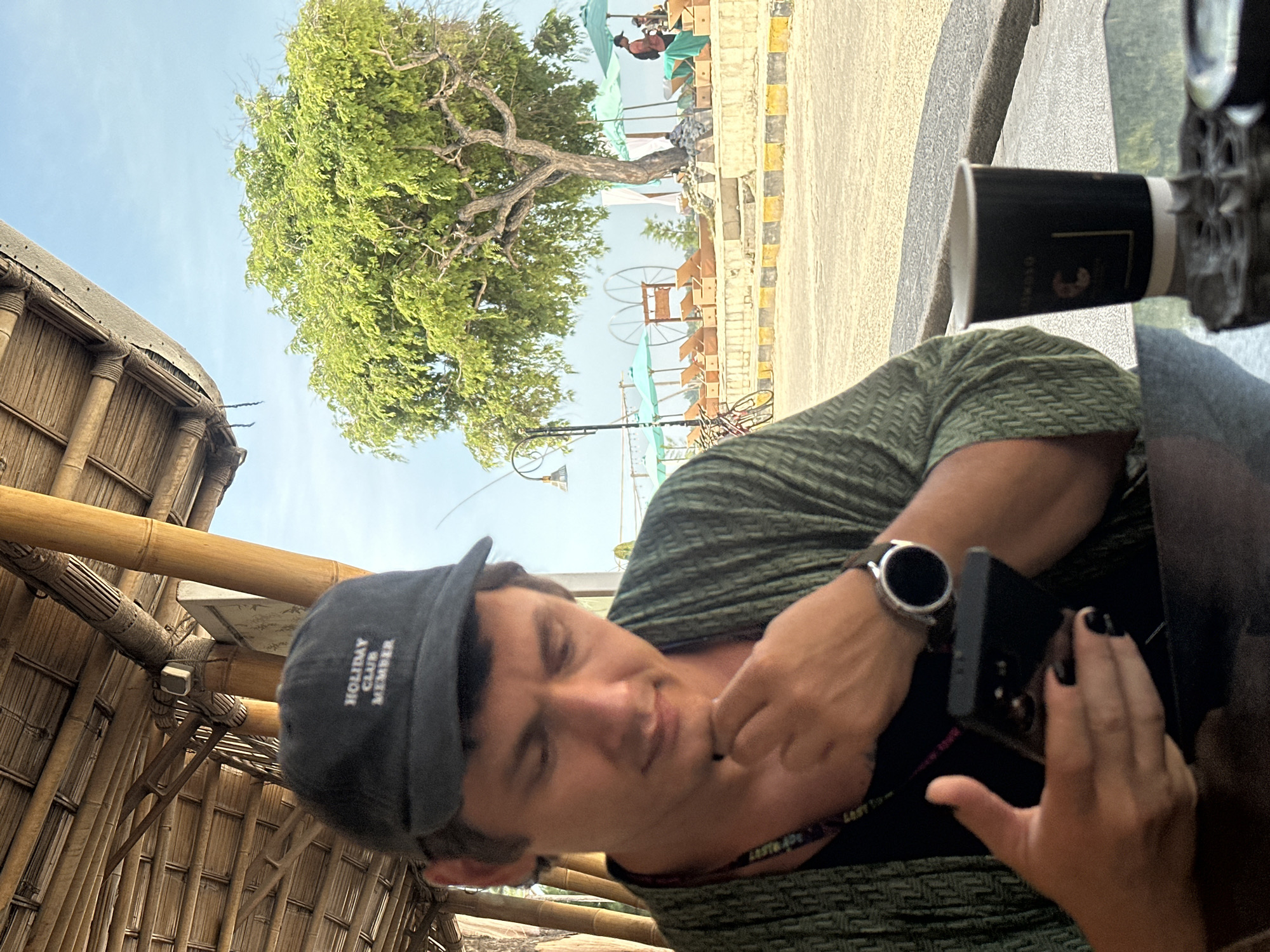 A man wearing a gray "HOLIDAY CLUB MEMBER" cap and a green patterned shirt is holding a black smartphone with painted black fingernails in his right hand and a smartwatch on his left wrist. He is sitting in a building with a bamboo roof and wall. Outside the building, there is a tree, a beach with lounge chairs and umbrellas, and a paved area. The location is in an unknown city in Indonesia.