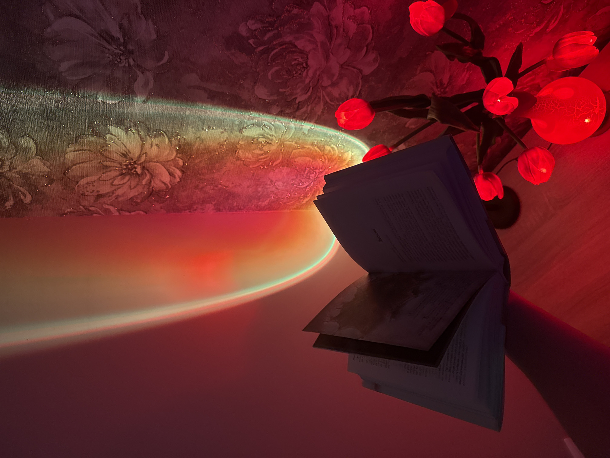 The image depicts an indoor scene bathed in warm, artificial lighting that casts vibrant red, orange, and green hues. In the foreground, an open book lies on its side, its pages filled with text and some illustrations. To the right of the book, a lamp shaped like a cluster of red tulips, with a round, cracked base, emits a strong red light. The background features a textured wall with a floral pattern, partially illuminated by the colorful light. The overall impression is one of a cozy, atmospheric setting, possibly a room in a private residence. The location is noted as Unknown, Unknown.