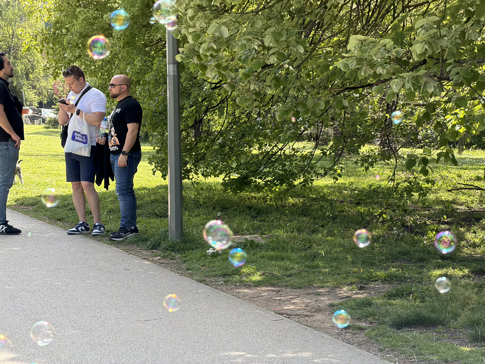 The image, taken on a sunny day, depicts a scene in a park. Three men stand on a paved walkway adjacent to a grassy area with trees. The first man, on the left, is wearing a black shirt and jeans, and faces away from the camera. The second man, closer to the center, is looking at a smartphone and is wearing a white polo shirt, blue shorts, and sunglasses. The third man, to the right, is wearing a black T-shirt, jeans, a watch, and sunglasses. Bubbles are scattered throughout the image, some near the people and others in the grassy area. The park appears to be well-maintained with lush greenery. Given the style of the buildings in the background and other details, the location is likely somewhere in the city of Sofia, Bulgaria.