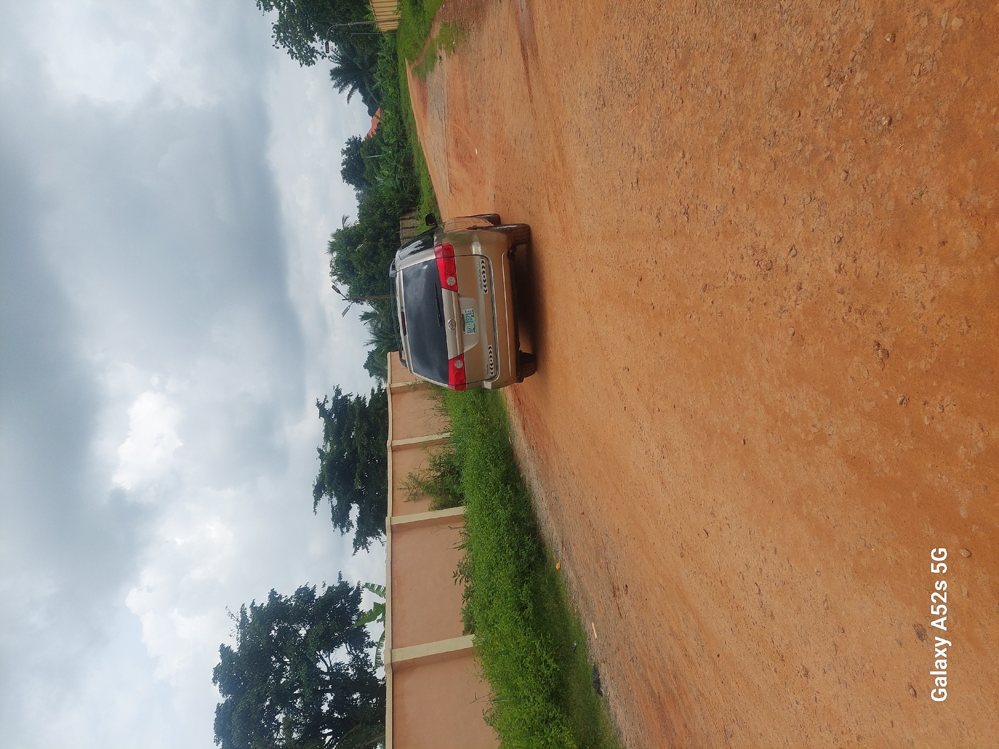 The image depicts a dirt road in Umudioka, Nigeria, with a beige minivan parked on the right side of the road, facing away from the viewer. The road is unpaved and appears dry, with scattered small stones and pebbles. To the left of the road, there is a low, light brown wall with white vertical supports, and beyond the wall, lush green vegetation and trees are visible. In the background, more trees and foliage line the road as it curves to the right, leading towards a residential area with a glimpse of orange-tiled roofs. The sky is overcast with heavy grey clouds. The watermark "Galaxy A52s 5G" is present in the bottom left corner of the image.