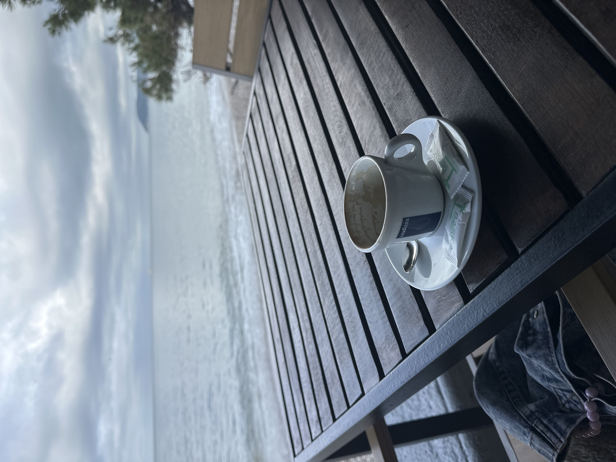 An empty coffee cup on a saucer sits on a dark, slatted wooden table, overlooking a calm sea under a cloudy sky. The image is taken from a slightly elevated perspective, looking down at the table and out towards the horizon.
In the foreground, the white ceramic coffee cup is empty, with some residue visible inside. It features the "LAVAZZA" logo on its side. On the saucer are two white sugar packets with green branding; one clearly shows the word "ZUCCHERO" (Italian for sugar). A metal spoon also rests on the saucer. The table has a robust, dark wood construction with visible grain and slats. Partially visible beneath the table on the right is a dark denim jacket draped over a chair or railing, alongside a pale pink beaded accessory.
The background reveals a vast, serene body of water, likely the sea, with gentle ripples on its surface. Above the water, a soft, overcast sky dominates, characterized by diffuse light and light grey clouds. In the upper left corner, dark green foliage, possibly pine needles, hangs into the frame, suggesting nearby trees. The overall atmosphere is quiet and reflective, indicative of a peaceful moment. This scene is consistent with a coastal setting, such as Šušanj, Montenegro, where one might enjoy a coffee overlooking the Adriatic Sea.