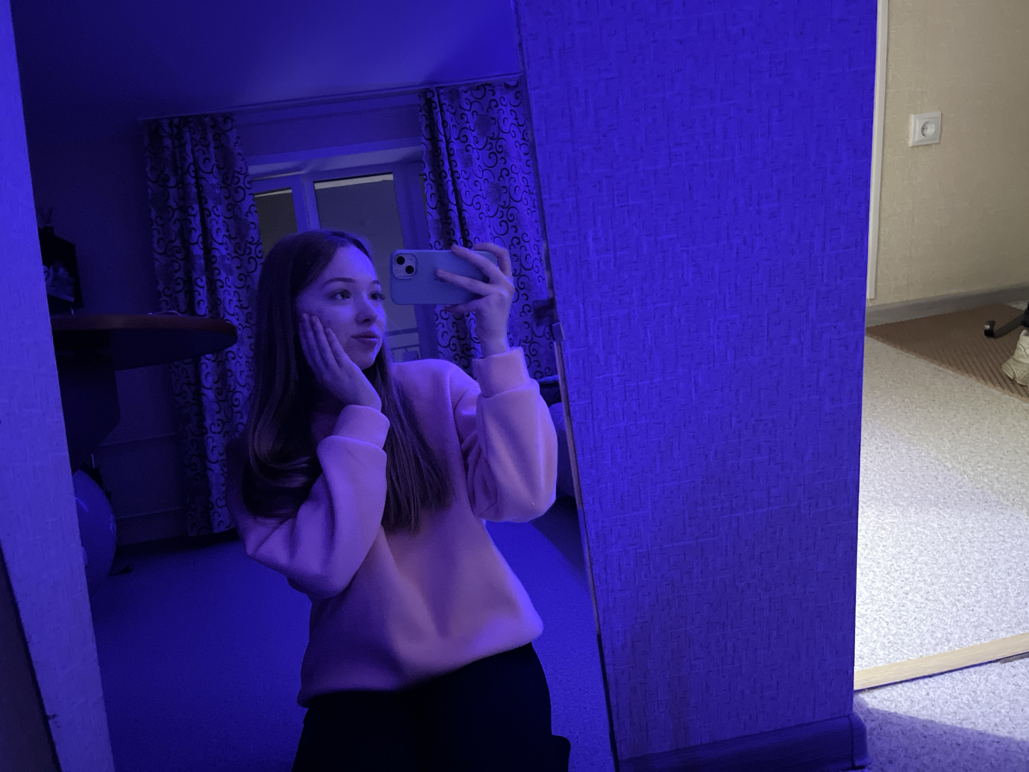 A young woman is taking a selfie in a mirror. The room is lit with blue light. The woman is wearing a light pink sweater and dark pants. She is holding a light blue phone in her right hand and resting her left hand on her face. Behind her is a window with patterned curtains. A power outlet is visible on the wall to the right of the mirror. The location is in an apartment, presumably in the city of Chaikovsky, Russia.