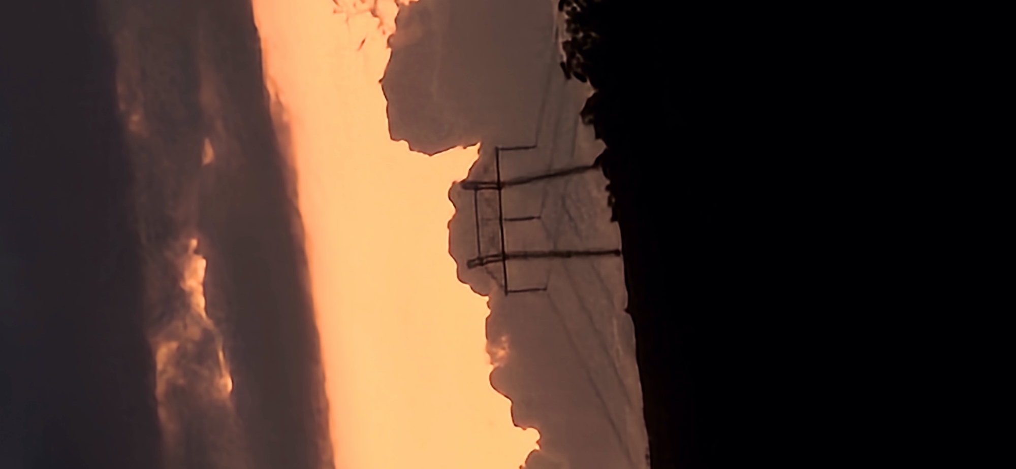 The scene, set in the city of Gutsal, Bulgaria, depicts a twilight landscape during sunset or sunrise. The sky is divided into distinct bands: the upper portion is dominated by dark, heavy clouds in deep gray and subtle purple tones, with minimal light penetration. Below this, a wide, vibrant horizontal band of bright orange and golden light extends across the mid-ground, indicating the low position of the sun. Interspersed within this brighter region are silhouetted, irregularly shaped clouds, some with illuminated edges.
A prominent electricity transmission pylon stands slightly to the right of the frame's center, rendered in a dark silhouette against the luminous orange sky. Multiple power lines extend horizontally from the pylon, visible as thin, dark lines against the brighter background. The lower third of the image consists of a completely dark, featureless silhouette of the ground line or distant hills, with some irregular, textural protrusions on the right suggesting sparse trees or vegetation. No people, animals, or explicit actions are discernible.