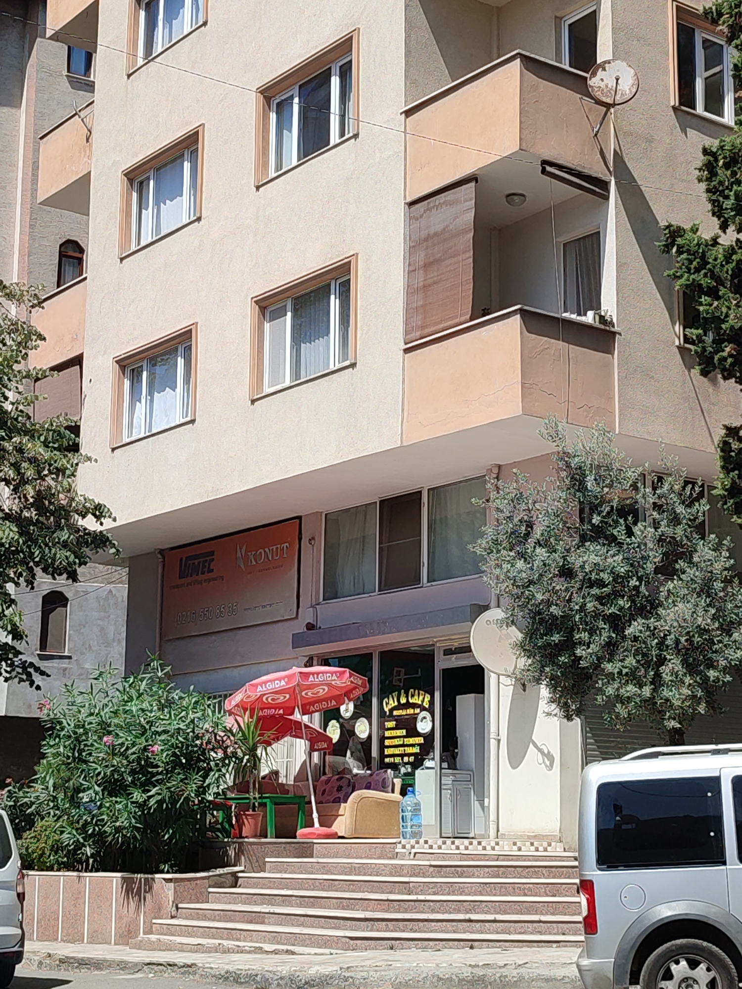 The image captures a street-level view of a commercial building in Kadıköy, Turkey. The building features multiple floors with cream-colored walls and brown-trimmed windows. A satellite dish is mounted on a balcony on the third floor. On the ground floor, a sign for "VINTEC KONUT" is visible, along with a telephone number. To the right, a cafe named "ÇAY & CAFE" is open for business, with red umbrellas providing shade outside. A silver van is parked on the street to the right of the cafe. Green bushes and trees are planted in front of the building, and steps lead up to the cafe entrance. The sky is bright, indicating daytime.