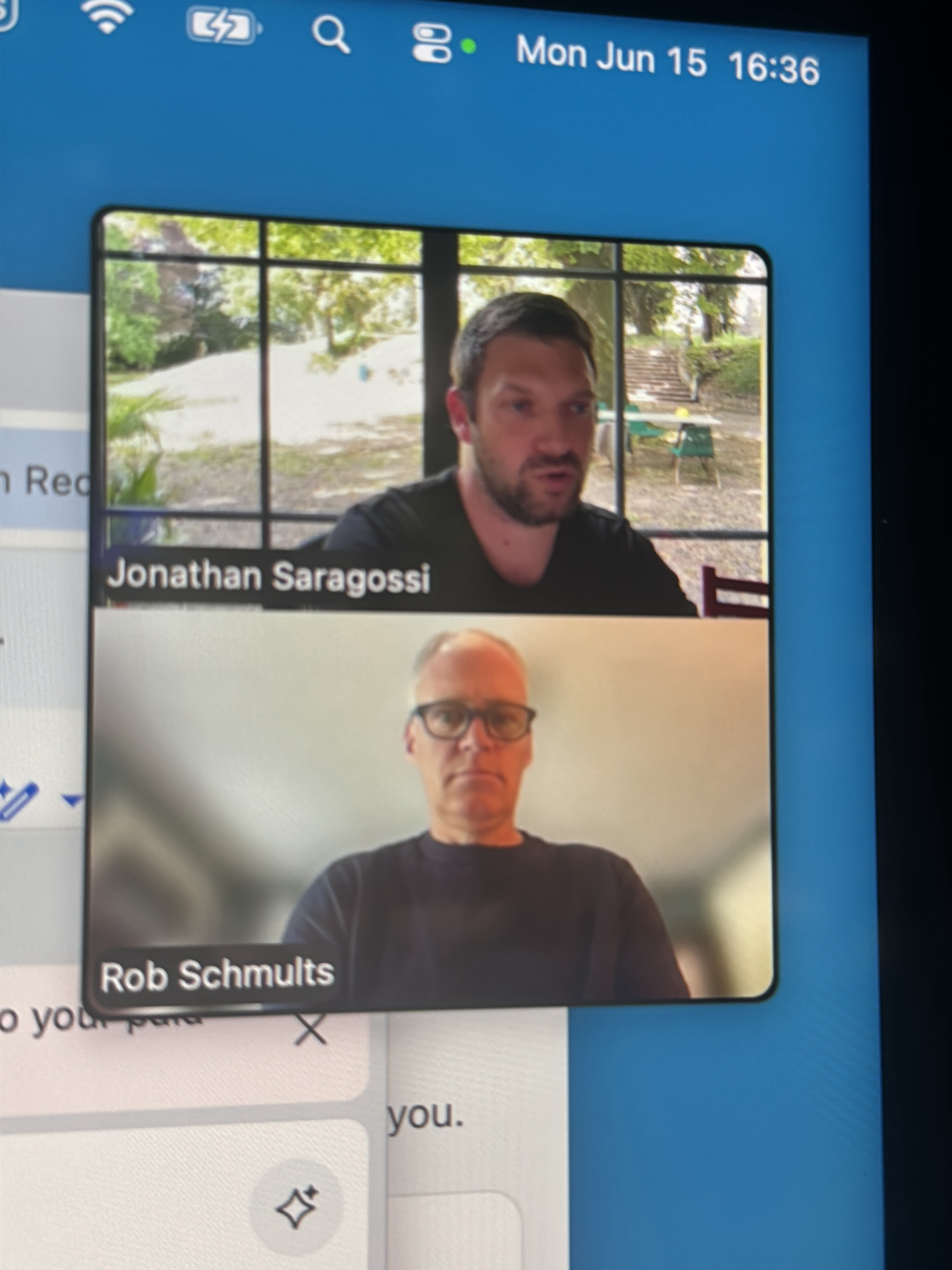 The image shows a screenshot of a video call interface. The call features two participants, Jonathan Saragossi and Rob Schmults, displayed in separate video windows. Jonathan Saragossi is in the upper window, facing forward and speaking. He has short brown hair and a beard and is wearing a black t-shirt. The background behind him appears to be an outdoor scene viewed through a large window, with trees and some outdoor furniture visible. Rob Schmults is in the lower window, facing forward and wearing glasses and a dark long-sleeved shirt. The background behind him is a blurred interior setting. The top of the screenshot shows system icons including a Wi-Fi signal, a battery indicator, and a search icon, along with the date and time "Mon Jun 15 16:36". Below the video windows, there is a text area with the partial text "o your para" and an "X" icon, suggesting a chat or annotation feature. The overall scene depicts a virtual meeting or conversation.