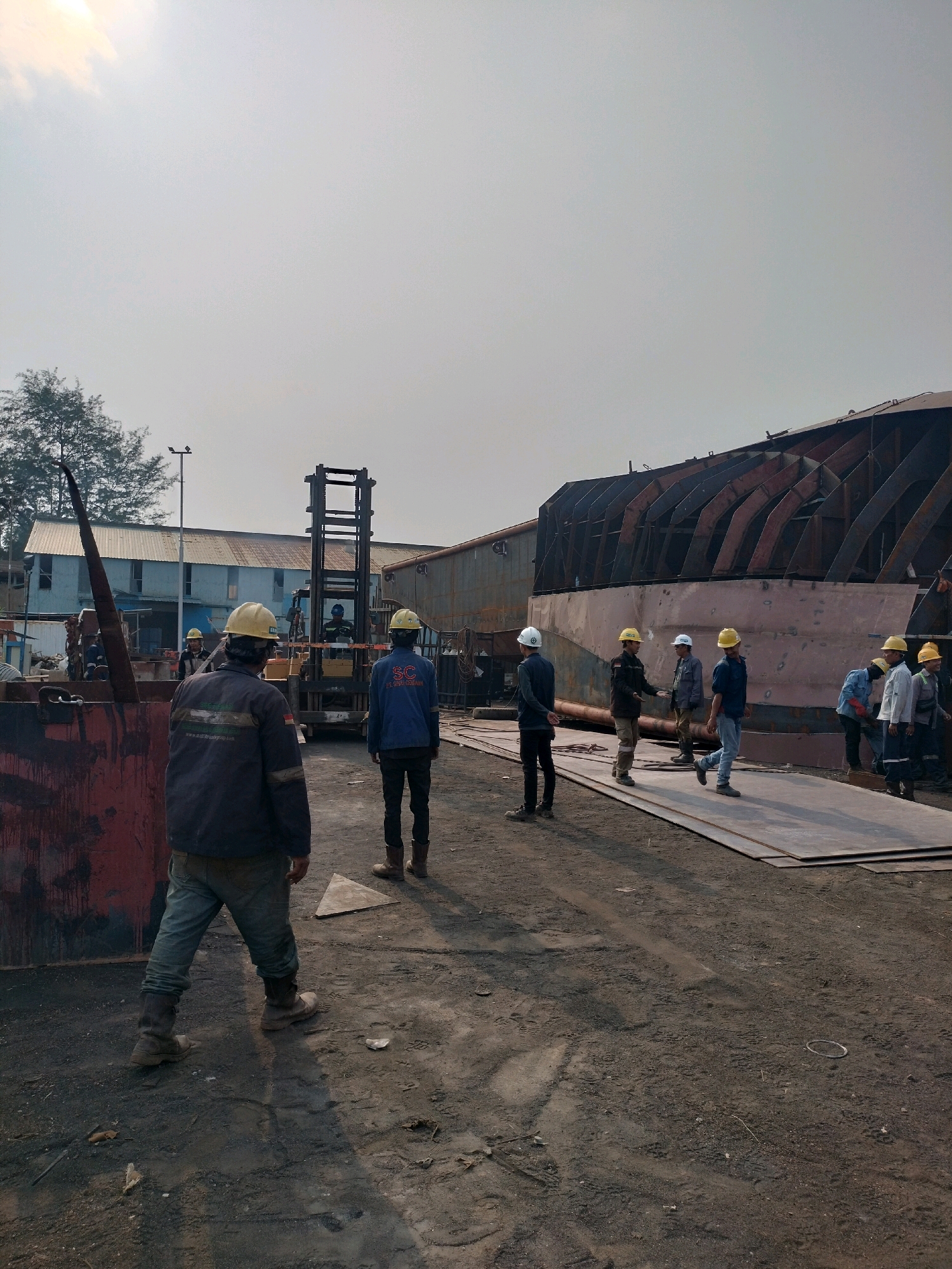 The image depicts an outdoor industrial site, likely a shipyard, in Batam, Indonesia. Several men, all wearing hard hats and work attire, are dispersed throughout the scene. In the foreground, a man in a dark jacket and light blue pants is seen from behind, facing away from the camera. To his right, another man in a blue jacket and black pants stands facing towards a large, partially constructed metallic structure, possibly a ship hull, which dominates the background.
Further back, a group of men are gathered near the metallic structure, some standing and observing, while others appear to be engaged in conversation or directing activity. A forklift is visible behind some of the individuals. The environment is dusty and unpaved, with scattered debris on the ground. The sky is overcast with a hazy sun visible in the upper left corner. The overall scene suggests an active work environment within a shipbuilding or heavy industrial facility.