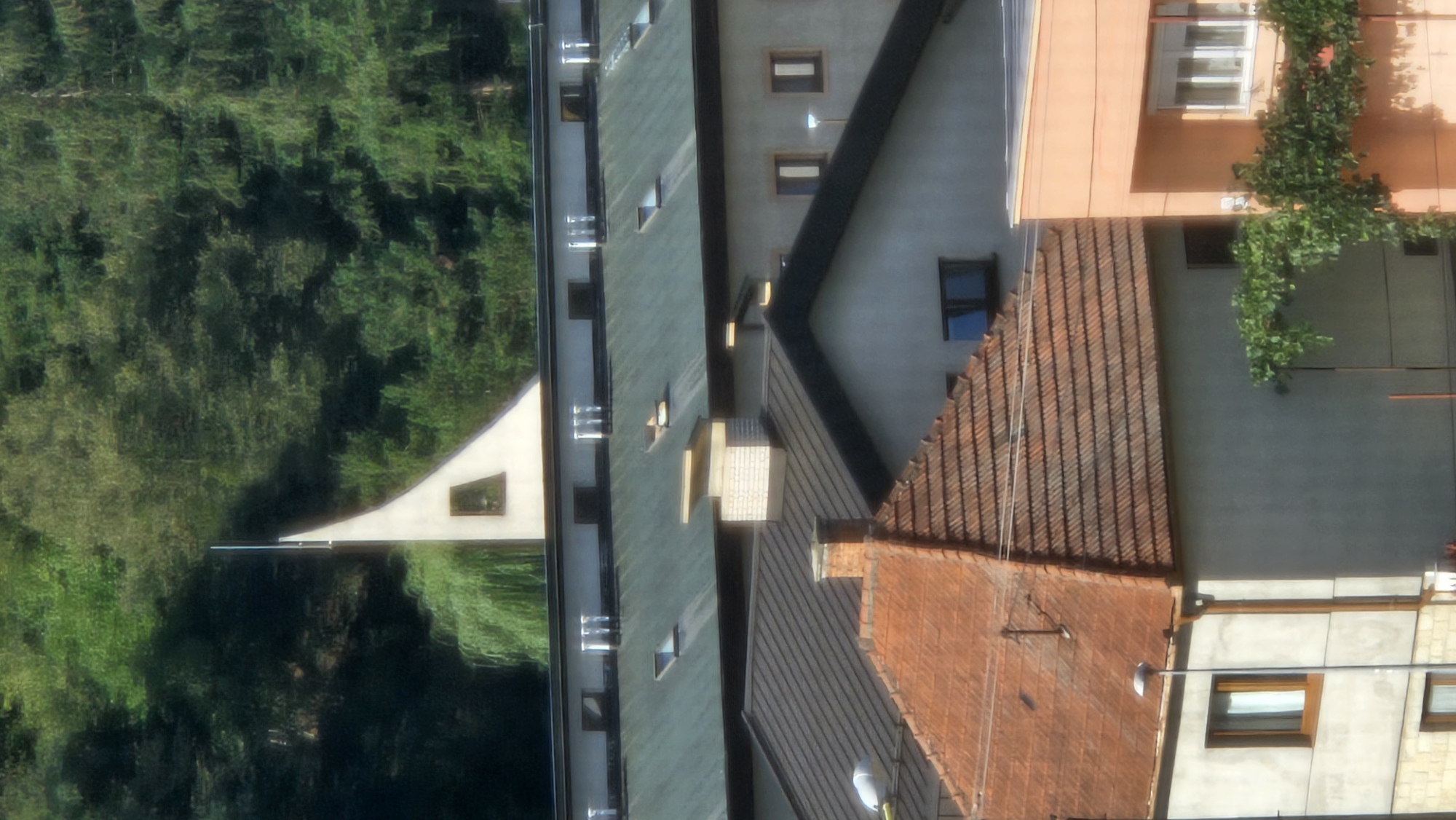 The image shows a densely wooded hillside with a modern white structure featuring a triangular roof and a narrow window. Below this, several buildings are visible. The closest structure has a prominent reddish-brown tiled roof and light gray walls, with several windows. To the right of this building, a section of an orange-clad building with white-framed windows is visible. In the middle ground, a long building with a dark green roof is adorned with multiple white exhaust vents or chimneys. Further back, a light gray building with dark-framed windows is visible, and above that, a row of smaller buildings with dark roofs and windows. The scene appears to be a residential or commercial area nestled against a natural, green backdrop. This vista is consistent with the hilly terrain surrounding Prešov, Slovakia, where modern architecture often intermingles with natural landscapes.