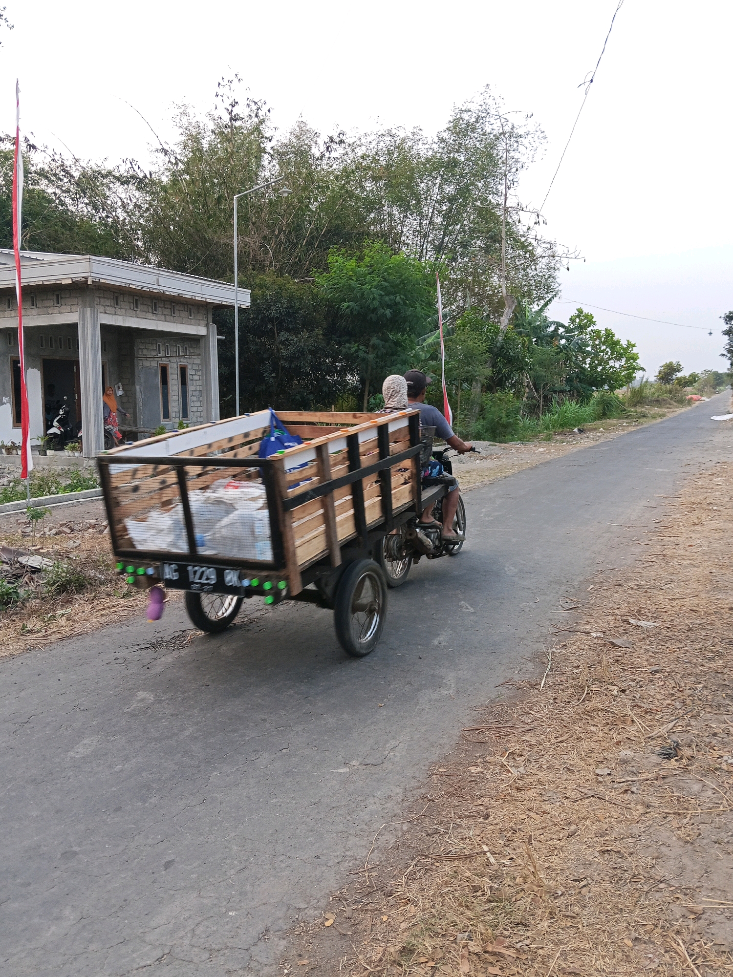 In Gogodeso, Indonesia, a man wearing a dark t-shirt and a black cap is operating a three-wheeled motorcycle that is towing a wooden trailer filled with various items. He is accompanied by a passenger wearing a light-colored headscarf and a dark-colored shirt, seated behind him. The motorcycle and trailer are traveling on an asphalt road lined with dry grass and dirt on the right side. On the left side of the road, a small, unfinished building with a metal roof is visible, adjacent to a flag pole with a red and white Indonesian flag. Trees and bamboo plants form a dense background. The sky is overcast. The trailer has a license plate that reads "AG 1229 BK".
