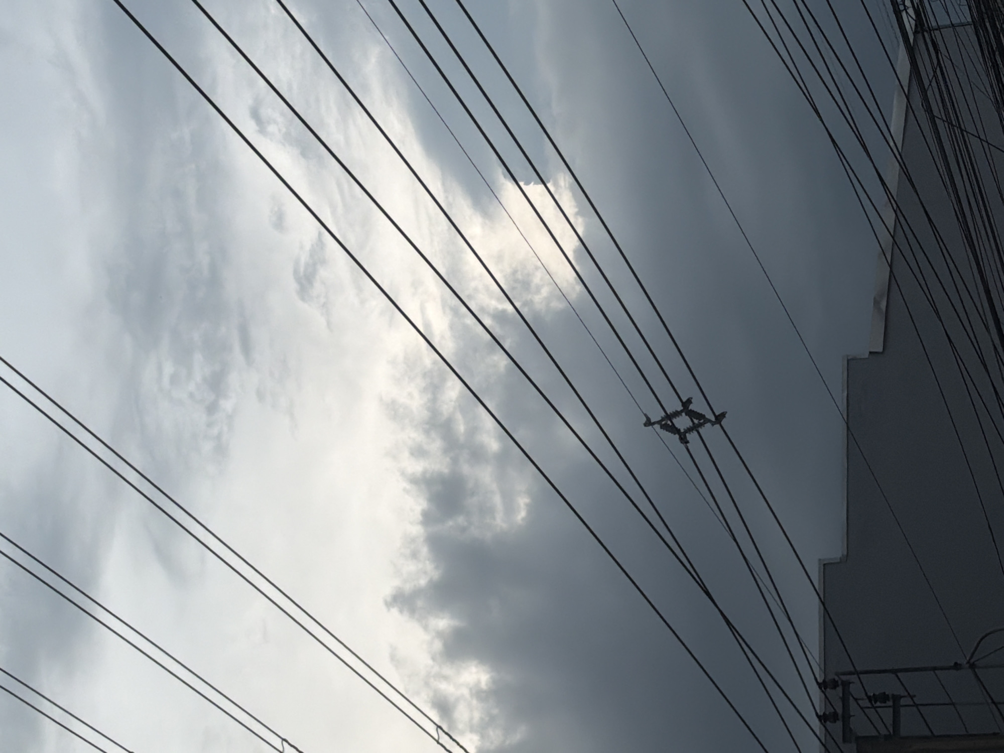 An overcast sky is visible in Bangkok, Thailand, filled with dramatic, dark clouds that obscure much of the sun. Numerous power lines crisscross the frame at various angles, creating a complex network against the muted sky. In the middle ground, a building with a light-colored facade is partially visible beneath the power lines. A dark gray, almost black, electric pole with insulators is also discernible on the lower left. The overall impression is one of a densely wired urban environment under a moody sky.
