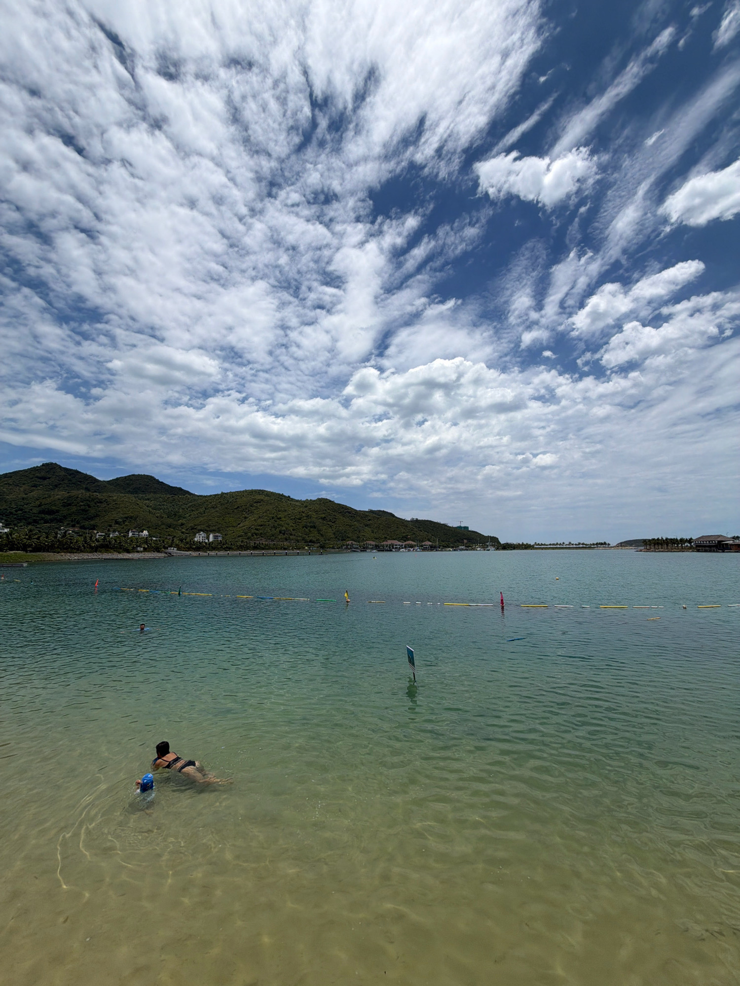The image shows a scenic view of the water and surrounding landscape in Nha Trang, Vietnam. In the foreground, two individuals are in the shallow water, one adult woman in a black swimsuit and a child wearing a blue swimming cap. The woman is lying on her stomach while the child is floating on their back. The water is clear and appears shallow near the shore, with sunlight creating ripples and patterns on the sandy bottom.
The middle ground features a calm body of water, likely a bay or lagoon, with a yellow and white buoy line demarcating an area of the water. Several buoys are visible, some red and some yellow, scattered across the water. In the distance, lush green hills covered in trees form the backdrop, with a few buildings visible nestled on the slopes. The sky is filled with a dramatic display of white, wispy clouds against a bright blue sky.
The overall scene conveys a sense of a peaceful and recreational day at a coastal location.
