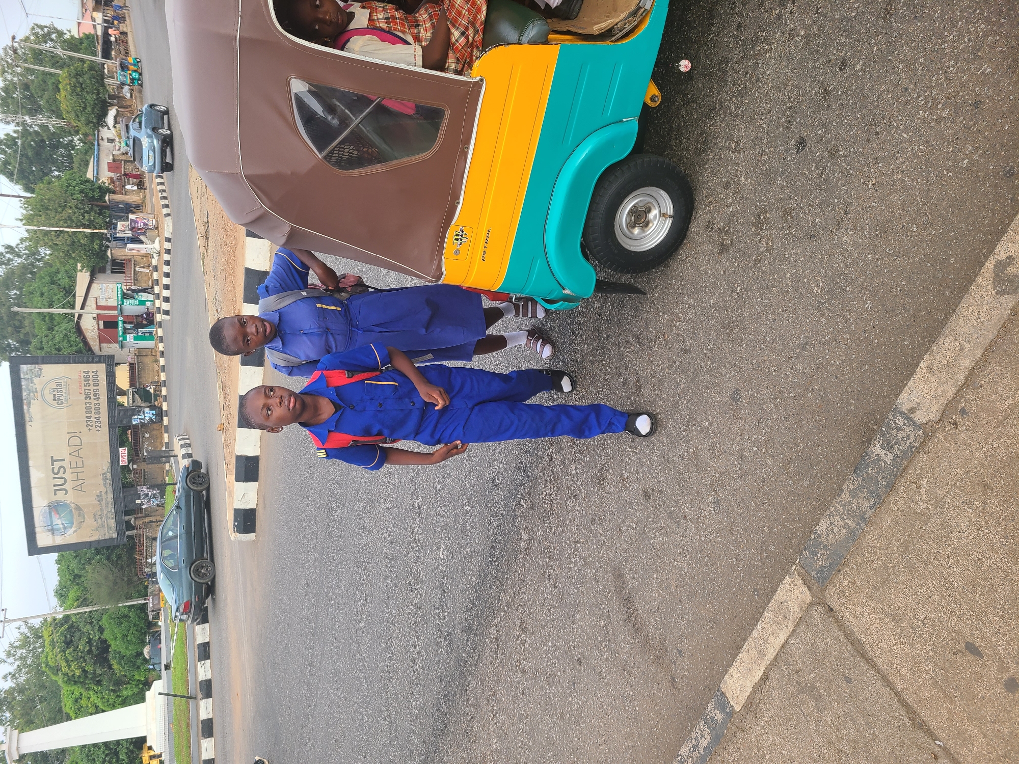 The image depicts a street scene in Jos, Nigeria, adjacent to a road. Two school children in matching blue uniforms stand next to a colorful 'keke' (a local motorized tricycle). The keke is painted brown, yellow, and teal. The road has black and white striped curbs. A billboard is visible, advertising "Crystal" water, with contact numbers starting with "+234". Other vehicles, including a dark-colored car, are visible on the road. Trees and buildings can be seen in the background. The road is paved with asphalt.