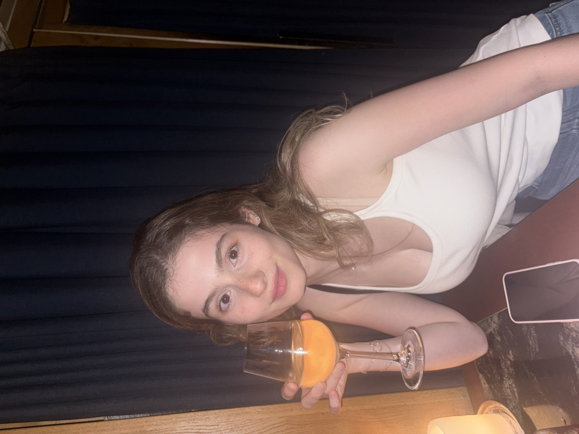 A young woman with fair skin and light brown hair is holding a wine glass filled with an orange liquid. She is wearing a white tank top and denim clothing. A mobile phone lies on the table. The background consists of dark blue curtains and wood panelling. The location of Tel Aviv, Israel cannot be determined from the image.