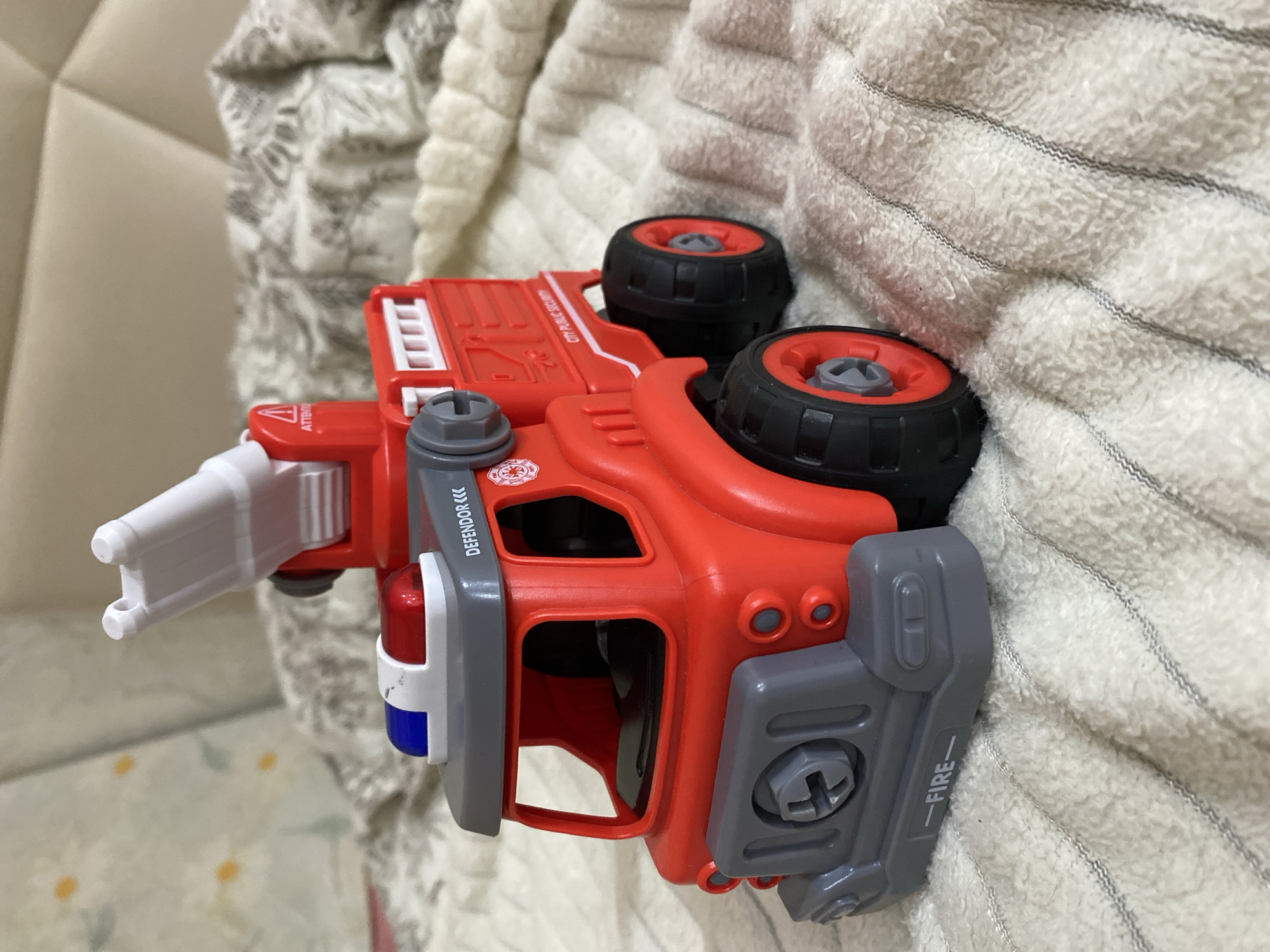 The image is a close-up, eye-level, indoor shot of a red toy fire truck. The fire truck is the primary subject and is positioned on a textured, cream-colored blanket. The blanket has faint, dark gray stripes running horizontally across it.
The fire truck is detailed, featuring a gray front bumper with the word "FIRE" printed on it, large black wheels with red rims, and a red body. It has a gray cabin with blue and red lights on top and a white ladder extended upwards. There are various labels and details on the truck, including "DEFENDOR" and a circular emblem with a fire symbol. The background is softly blurred, showing a quilted cream-colored surface, possibly a headboard or furniture. No people are visible in the image, and no specific location in Balashikha, Russia, can be identified. The scene is static and appears to be a still-life arrangement of the toy.