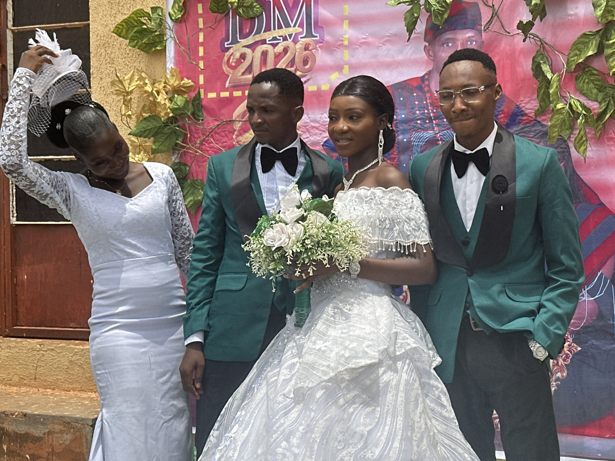 A group of four people stands in front of a backdrop for a wedding. The backdrop is pink and features green vines and a photograph of a man in a traditional headdress. Text on the backdrop reads "DM 2026." A woman in a long-sleeved white dress and a white fascinator is standing on the left. She is reaching up to adjust her fascinator with her right hand. Next to her is a man in a green suit with a black lapel and a black bow tie. A bride in a white wedding gown is in the center, holding a bouquet of white flowers. To the right of the bride is another man wearing a green suit with a black lapel and a black bow tie. This man is wearing glasses. The setting appears to be outdoors in the city of Unknown, Unknown.