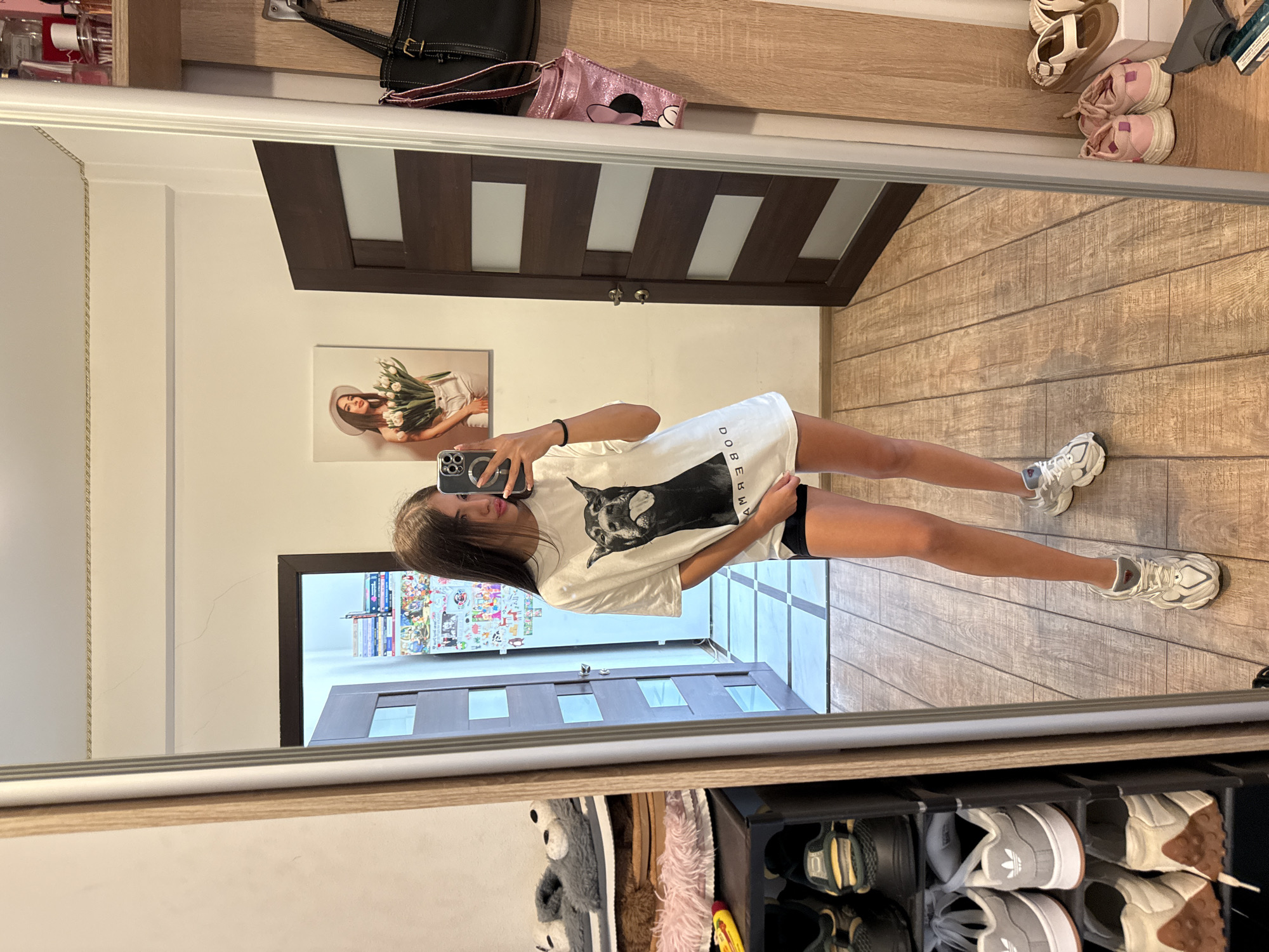 The image shows a young woman taking a mirror selfie in what appears to be a hallway or entryway area within a residential setting. She is standing in front of a large mirror, wearing a white t-shirt with a black dog graphic and black shorts. On the left side of the image, a shoe rack is visible, holding various pairs of sneakers and slippers. To the right of the woman, a dark wooden door with frosted glass panels is present, and further to the right, a wooden shelf unit is mounted on the wall, holding handbags and perfume bottles. The floor is made of light-colored wood-look tiles. A framed picture of a woman holding tulips hangs on the wall behind her. The scene is well-lit, likely by indoor lighting. The setting does not provide any specific visual cues to determine its location within Chișinău, Moldova.