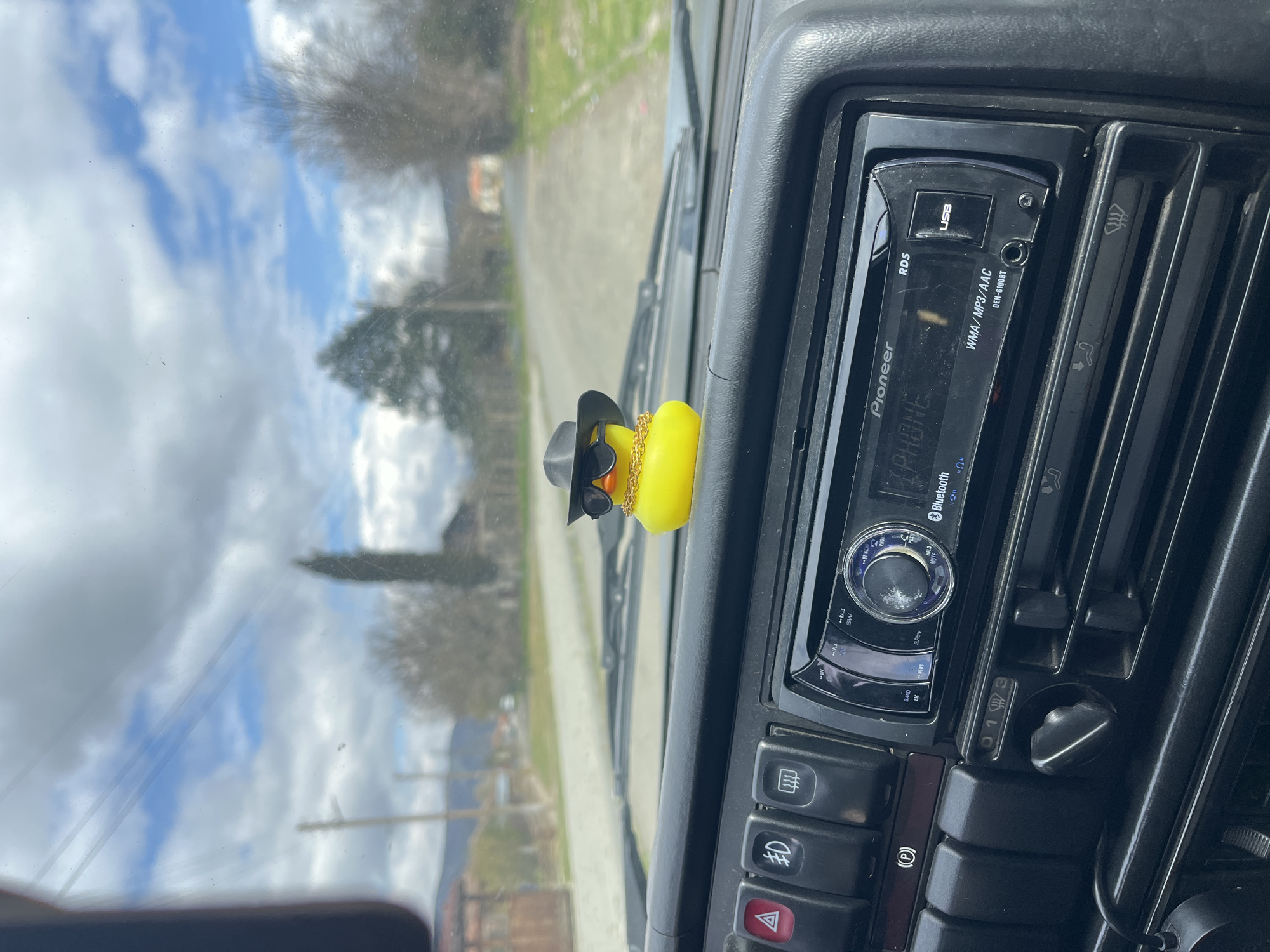 A view from inside a car in Lozarevo, Bulgaria. The dashboard features a Pioneer car stereo set to "iPhone" mode, along with various car controls and vents. On the dashboard, in front of the windshield, sits a yellow rubber duck wearing a black hat, sunglasses, and a gold chain. Outside, a blurred view of the road, trees, and buildings indicates the car is in motion. The sky is cloudy with patches of blue.