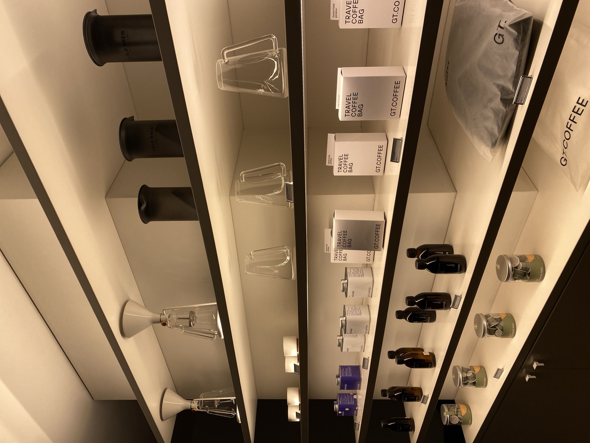 The image shows a display of coffee products in a store, possibly in Moscow, Russia. The products are neatly arranged on shelves and include items such as cold brew makers, glass mugs, travel coffee bags, coffee bottles, and coffee capsules. Some of the items are labeled in Russian, such as "Эфиопия ДЕМБИ" and "ГЕНИЯ КЕНИЯ," indicating that the coffee is from Ethiopia and Kenya.