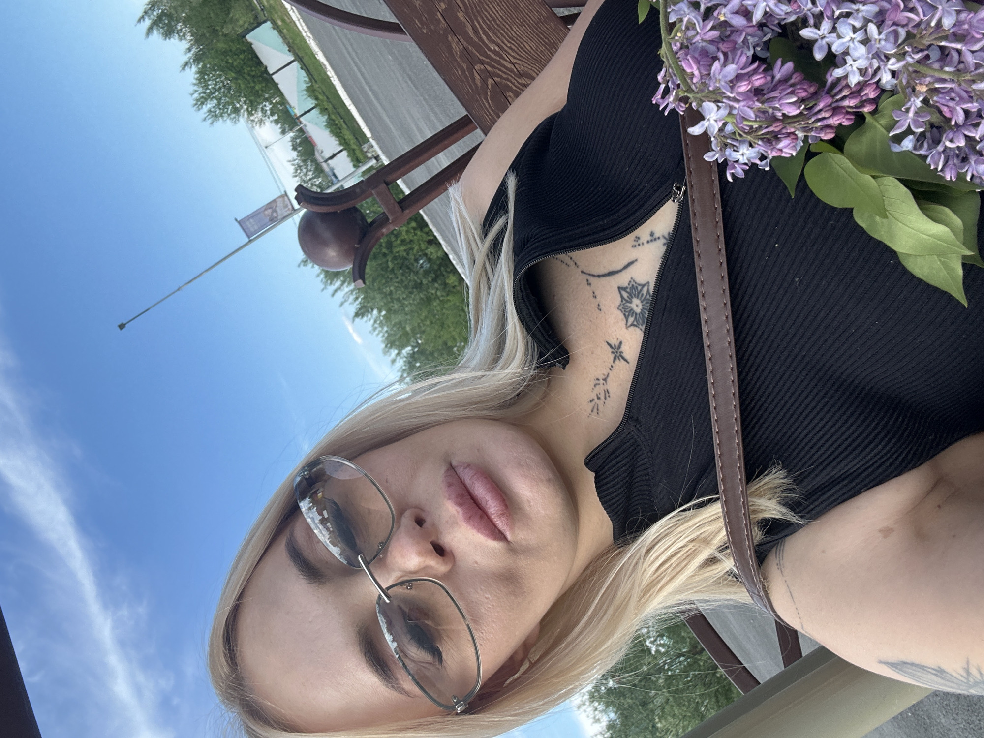 The image captures a blonde woman taking a selfie outdoors in the city of Unknown, Unknown. She appears to be in her 20s or 30s, looking directly at the camera through rectangular, rimless sunglasses with light-reflective lenses. Her lips are full and coated with a pinkish gloss. Tattoos are visible on her chest and collarbone area, featuring a geometric star-like design, various dots, and linear patterns. A partial tattoo resembling a feather or leaf is visible on her left upper arm. She wears a black, ribbed, high-necked top with a zipper, which is partially unzipped. A brown leather strap, possibly from a bag, crosses her chest. In the lower right foreground, she holds a bouquet of purple lilac flowers with green leaves. Behind her right shoulder, a dark brown wooden bench with a spherical decorative finial is visible. The background shows a clear blue sky with some white wispy clouds, green trees, a paved area, and a white and light blue patterned fence or wall. A tall, slender pole with a banner attached stands in the mid-distance. The scene is brightly lit by daylight, indicating an outdoor public or semi-public setting.