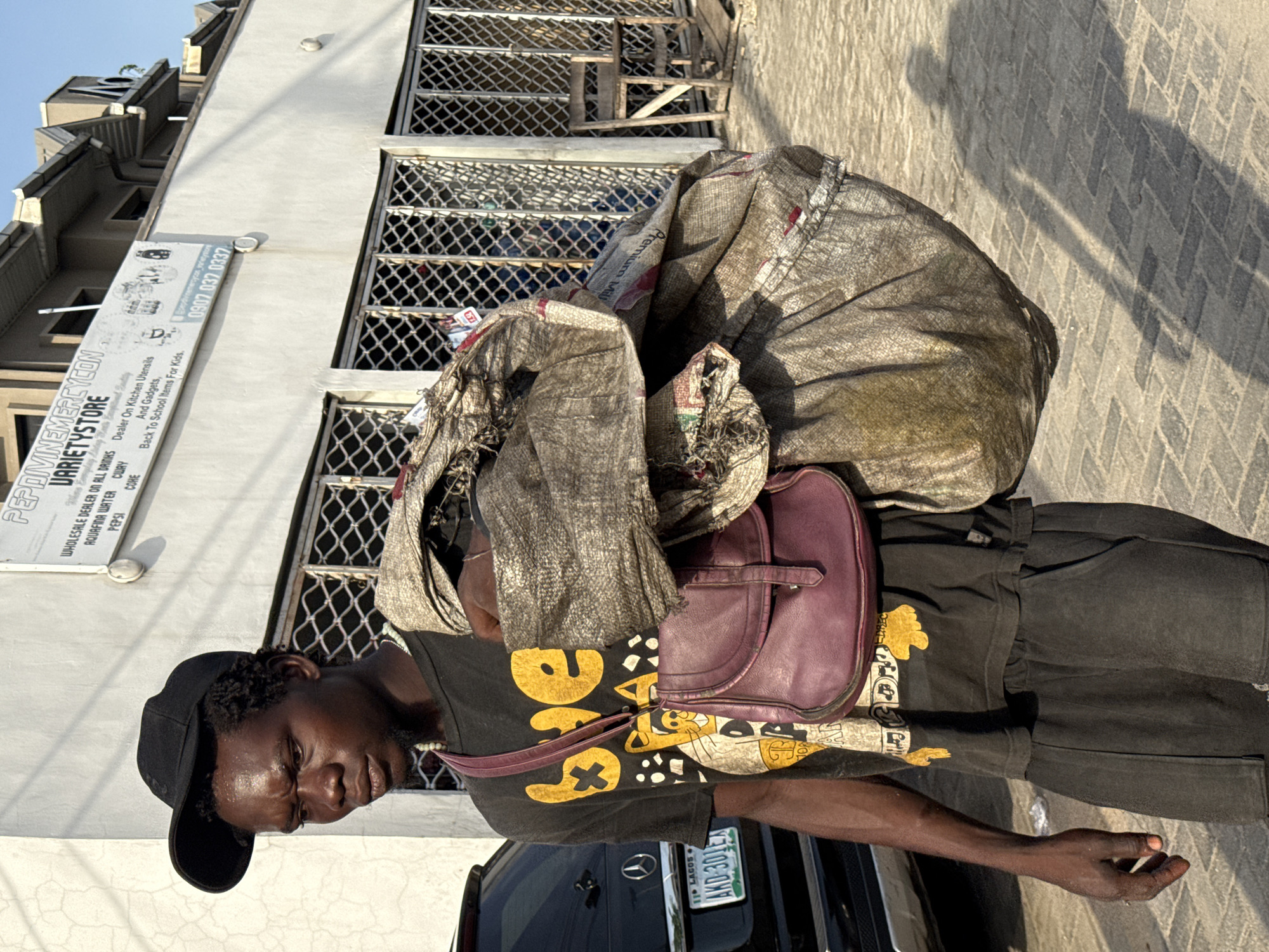 A man wearing a black T-shirt, black shorts, and a black cap carries a large bundle wrapped in gray fabric on his back, with a purple bag visible as part of the bundle. He stands on a paved sidewalk near a black car displaying a license plate reading "AKD-301EA" and the state name "LAGOS." In the background, there is a light-colored building with a sign that reads "PEPDIVINEMERCYCON VARIETYSTORE," indicating that it is a wholesale dealer of drinks and kitchen utensils/gadgets. The sign also lists the phone number "0907 037 0337." The location is suggested by the sign and license plate to be Ado, Nigeria. The lighting suggests it is daytime.