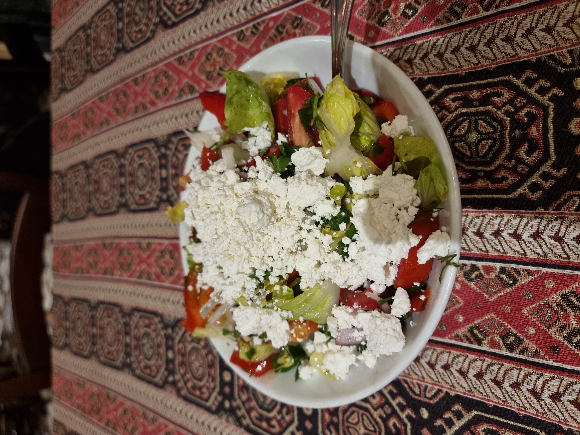 A close-up, slightly angled shot shows a white bowl filled with a salad covered in a generous amount of crumbled white cheese. The salad appears to contain lettuce, red bell peppers, and herbs. A fork is also visible in the bowl. The bowl is placed on a patterned tablecloth featuring a mix of red, black, and beige colors with geometric designs. The setting appears to be indoors, with part of a dark wooden chair visible in the lower left corner of the image. The image was taken in Givatayim, Israel.